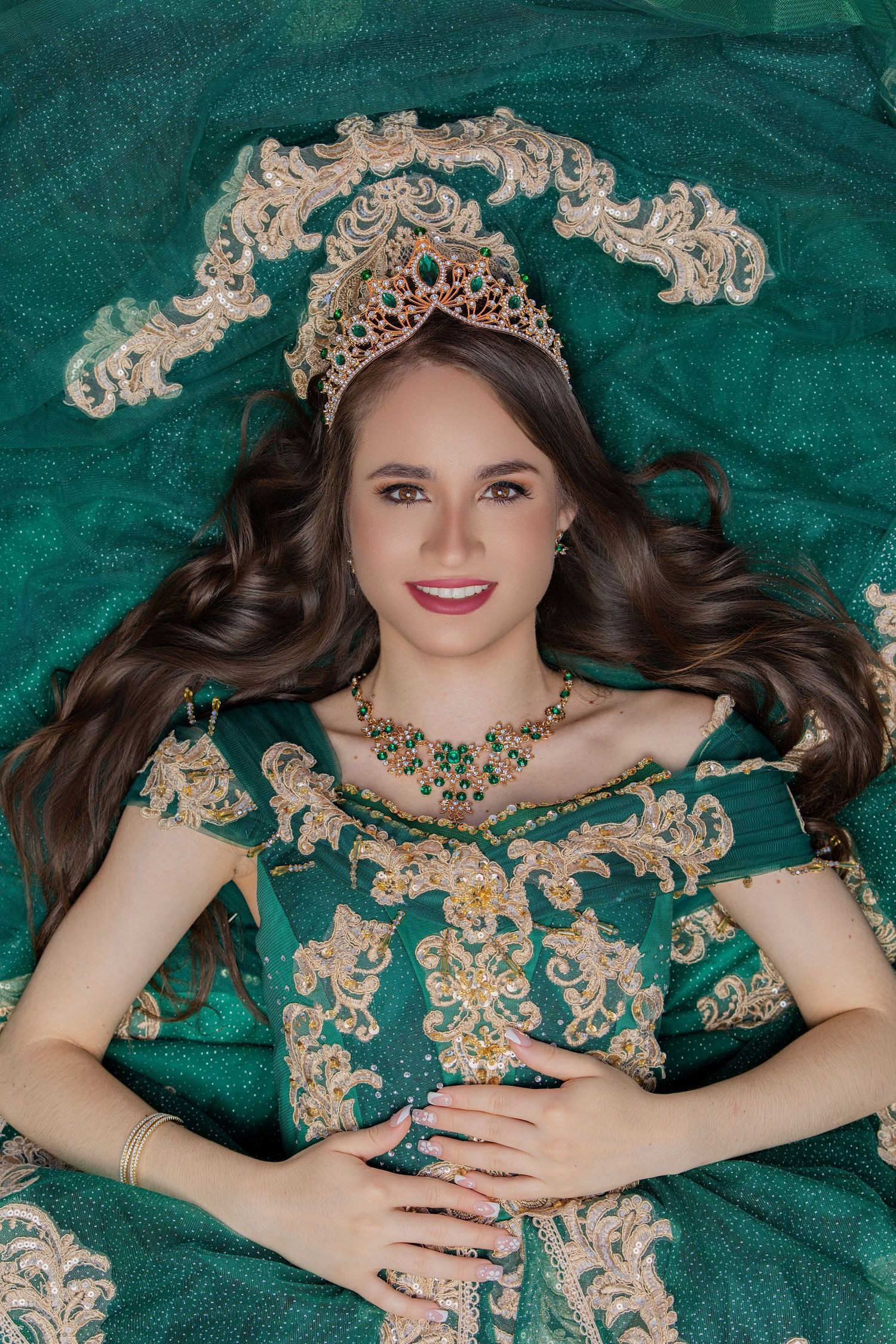 Young woman wearing an emerald green ballgown and crown lying down on a decorated backdrop