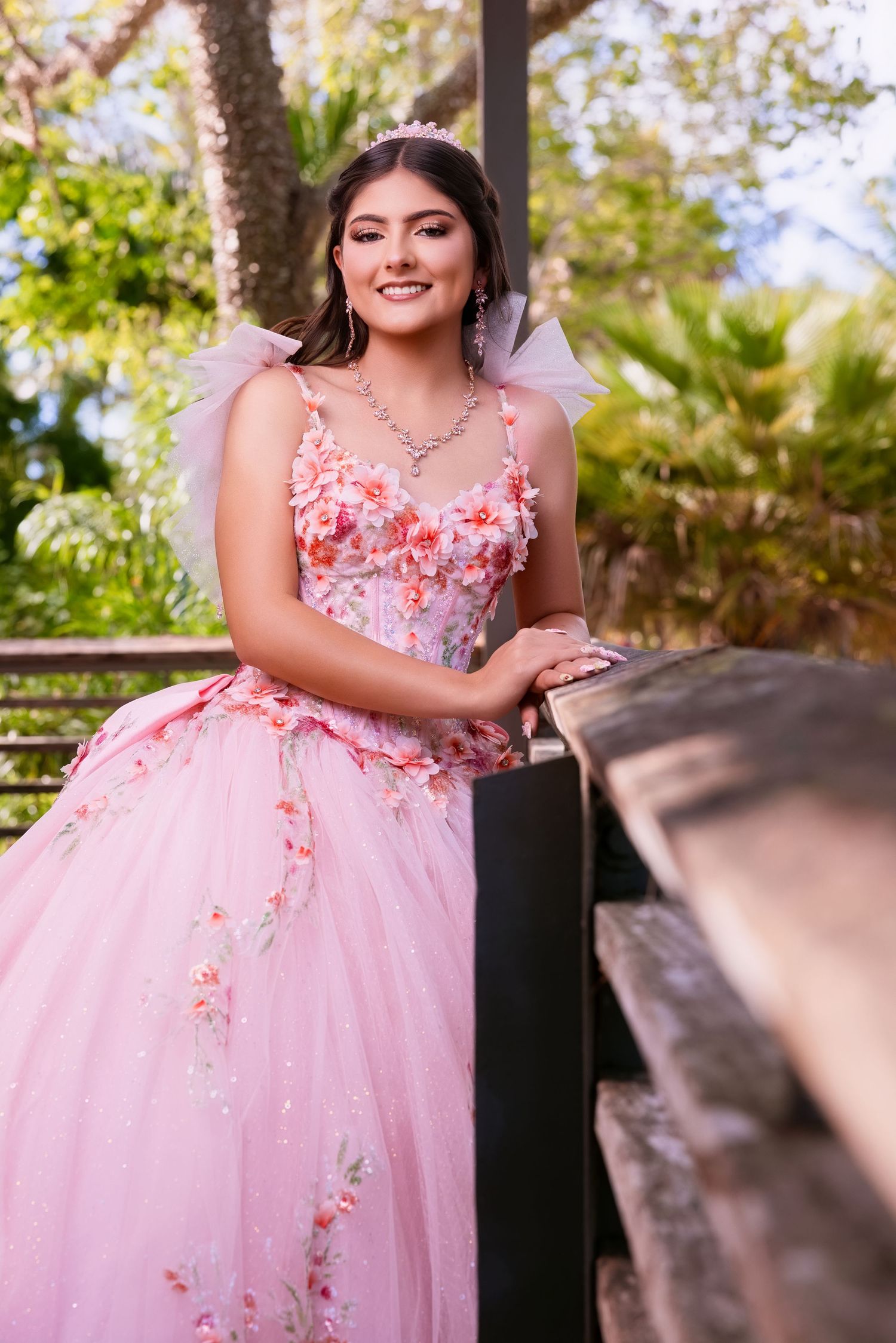 Smiling young woman wearing a pink strapless gown with floral embroidery posing outdoors next to a railing