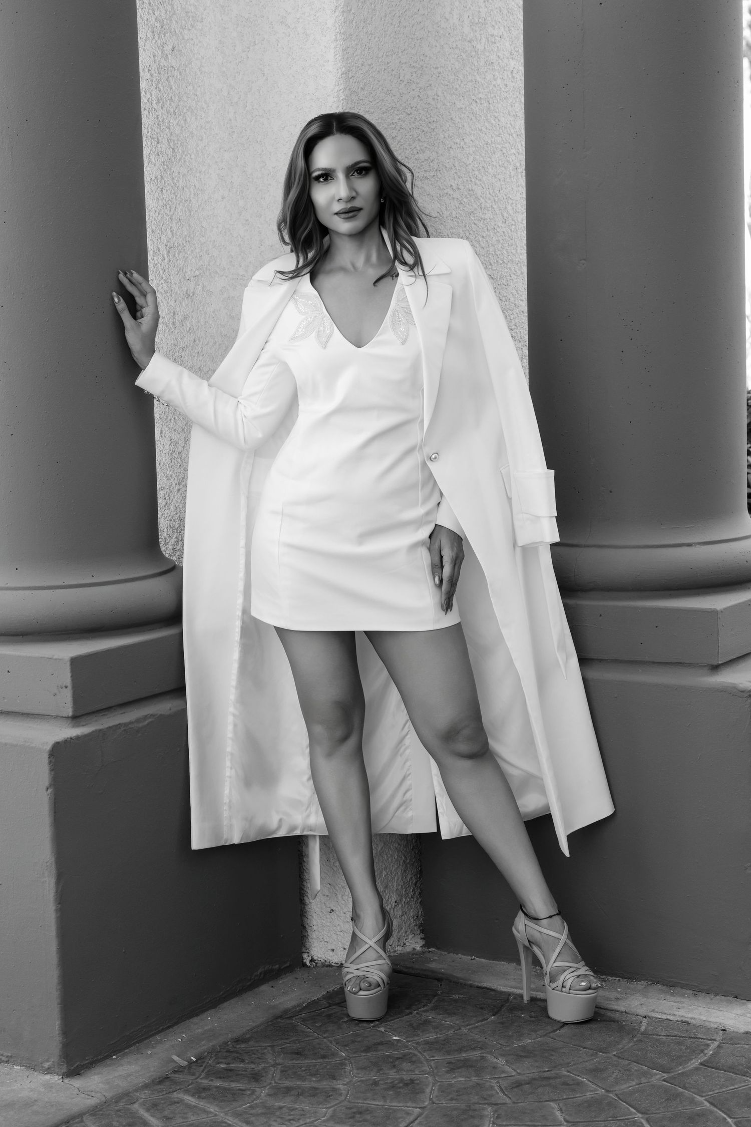 Full-length black and white fashion photography of a woman wearing a light mini dress and draped trench blazer leaning against a pillar.