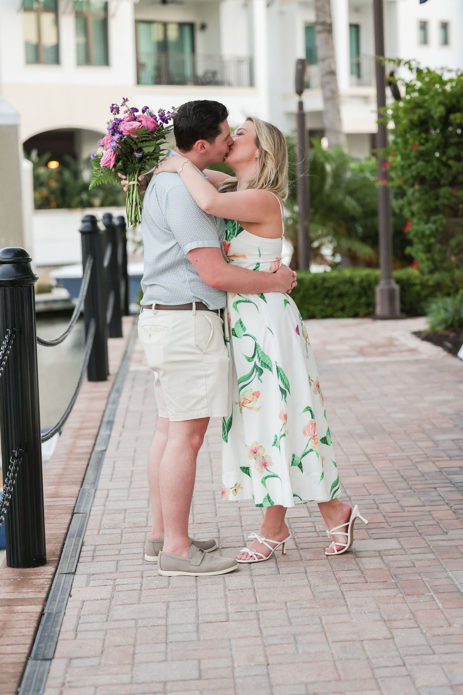 Couple kissing on a brick pathway outdoors, woman wearing a white floral midi dress