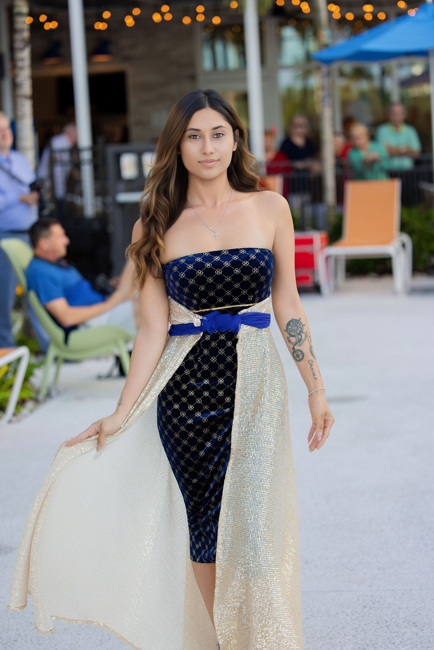 Woman walking outdoors in a strapless black mini dress featuring a long flowing tulle overlay