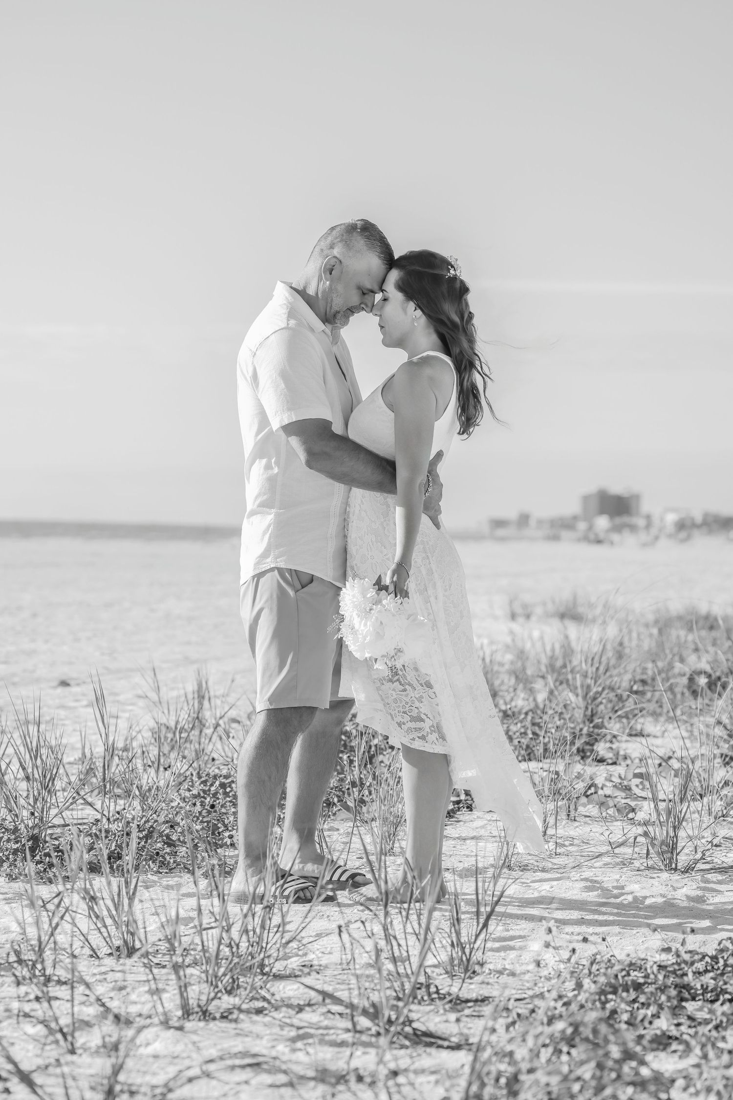 Black and white photo of a couple hugging outdoors on a beach with light vegetation