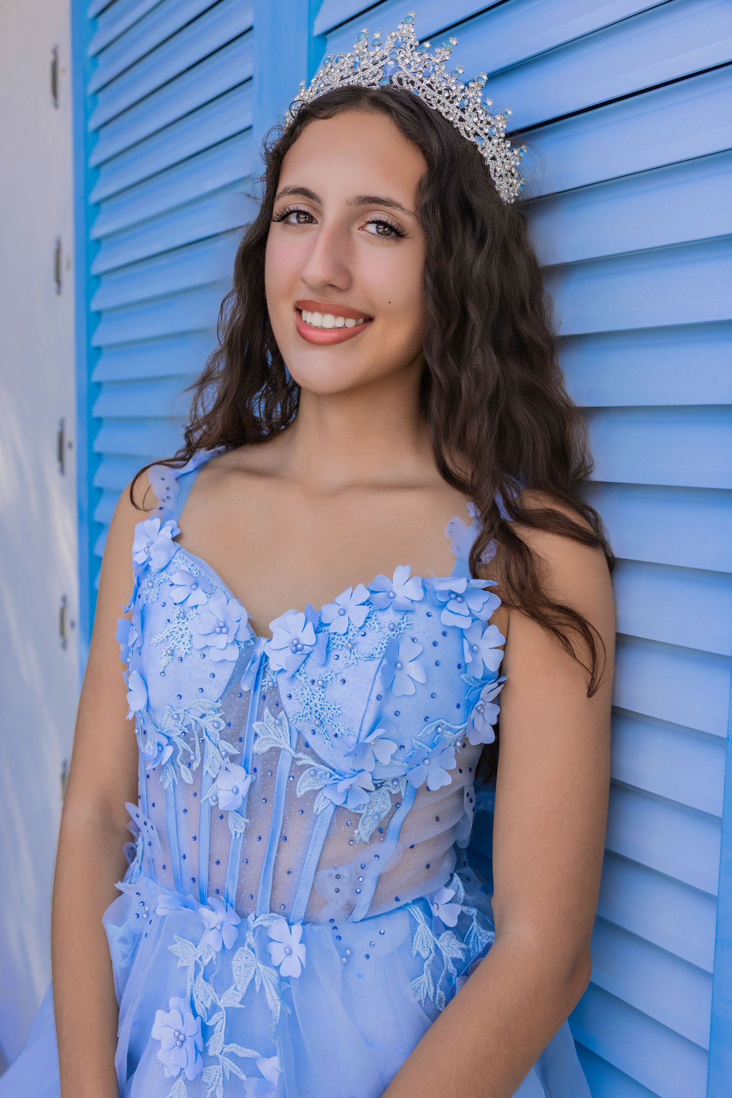 Smiling young woman wearing a blue floral embroidered dress and a tiara against blue shutters