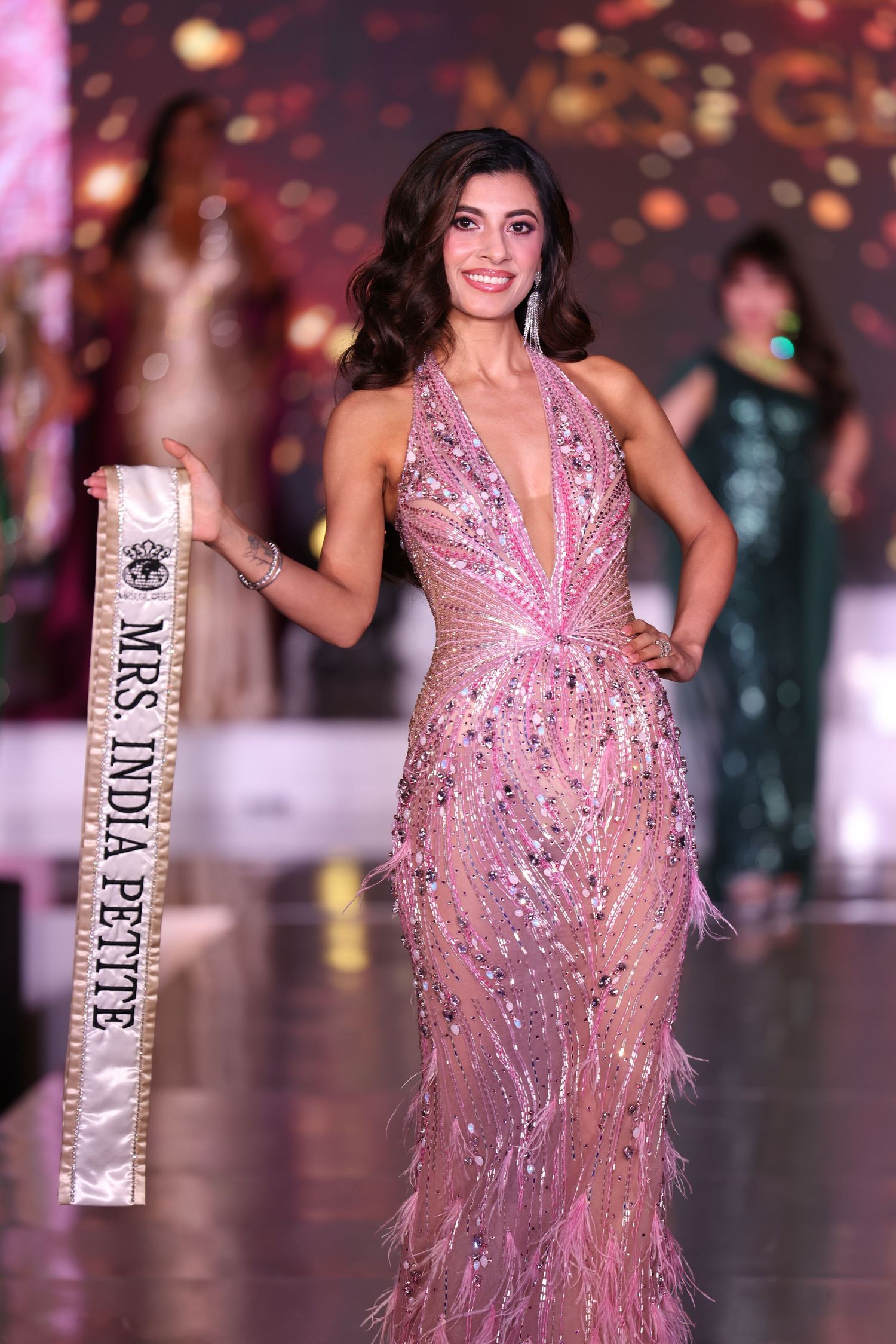 Mrs. India Petite Globe wearing an elegant evening gown on stage, holding her sash and looking toward the audience during a pageant event.