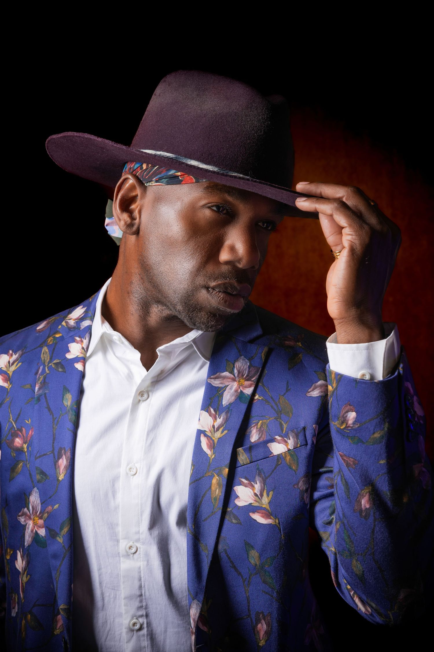 Man wearing a textured blue blazer and a wide-brimmed fedora hat touching the brim for a studio portrait.