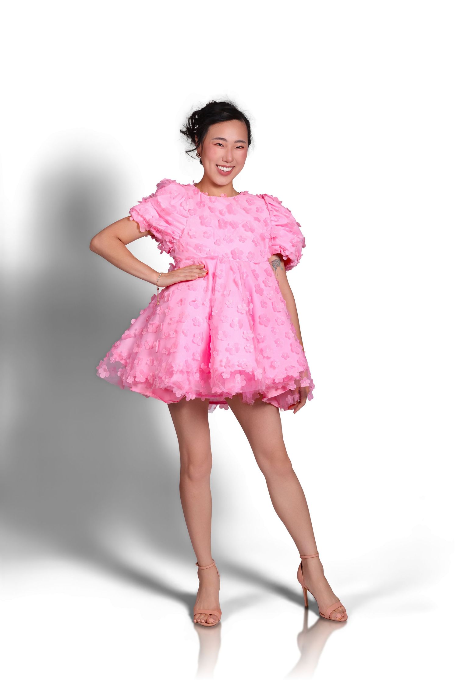 A woman modeling a voluminous, bright pink ruffled tulle mini dress with short puff sleeves against a plain white background.