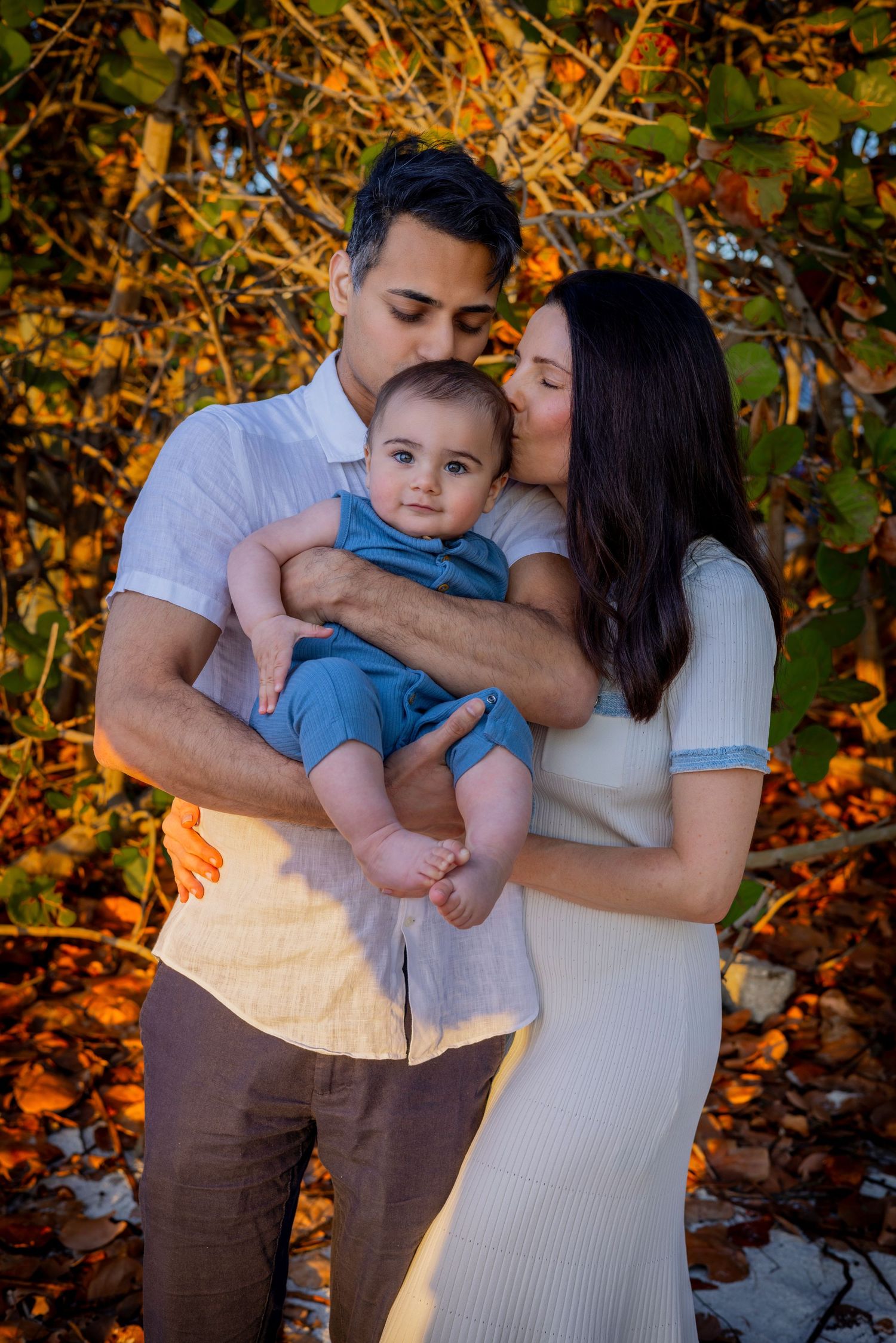 Parents hugging and smiling with their baby boy outdoors in a sunny garden setting