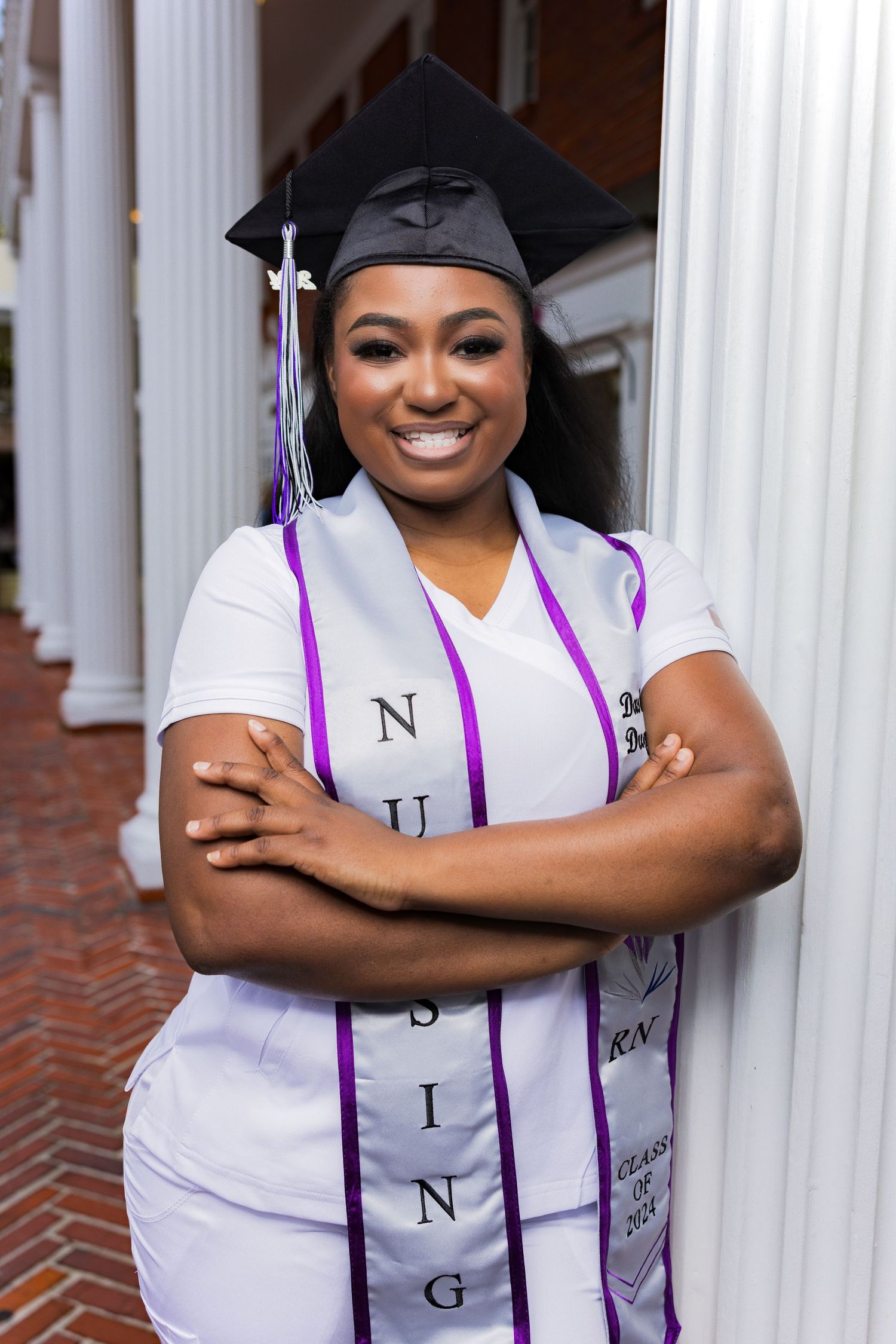 Smiling graduate in a white dress, black mortarboard cap, and purple academic stole leaning against a white pillarImage.
