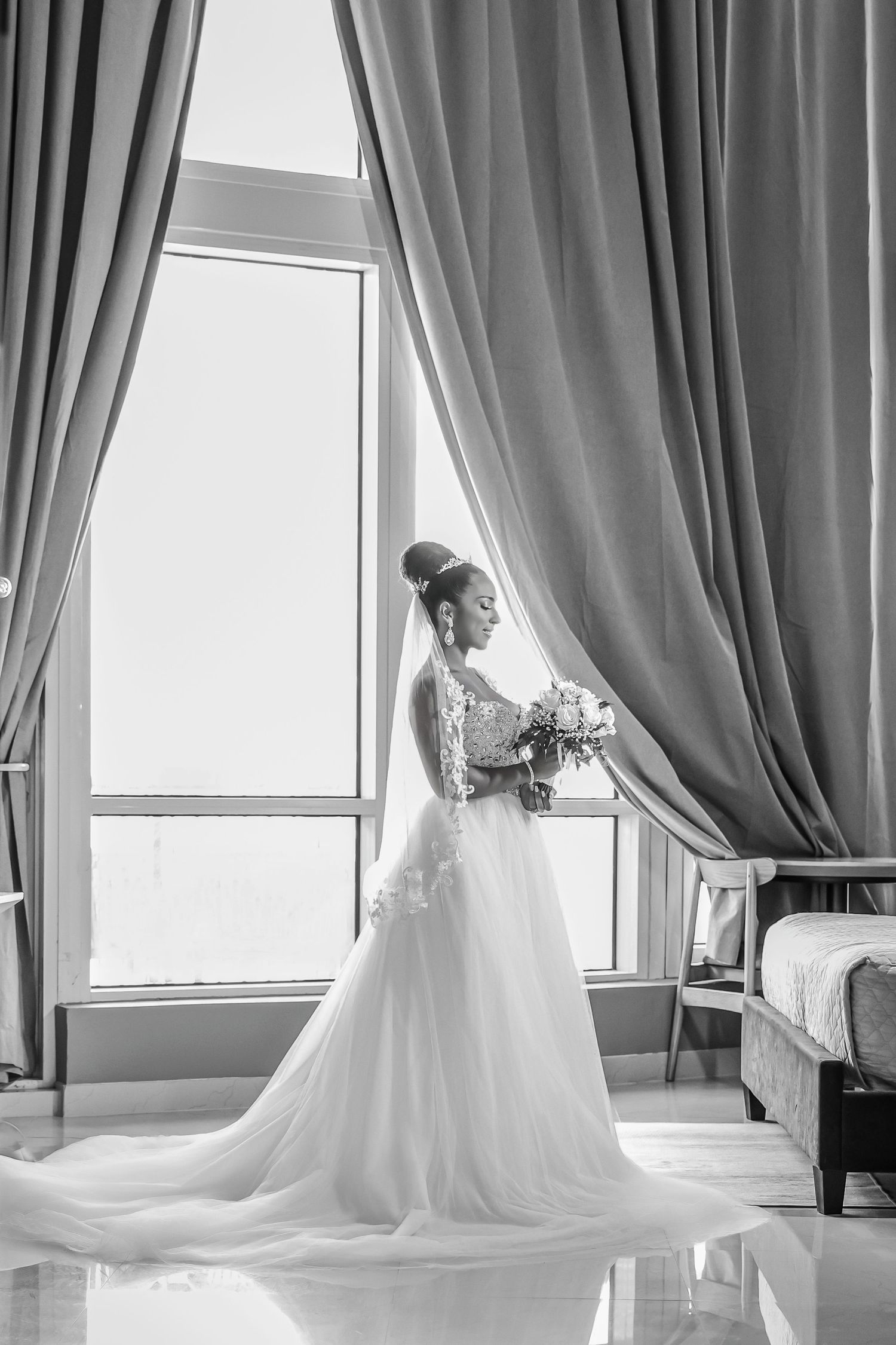 Black and white silhouette of a bride in a full ballgown looking out a large window framed by drapes.
