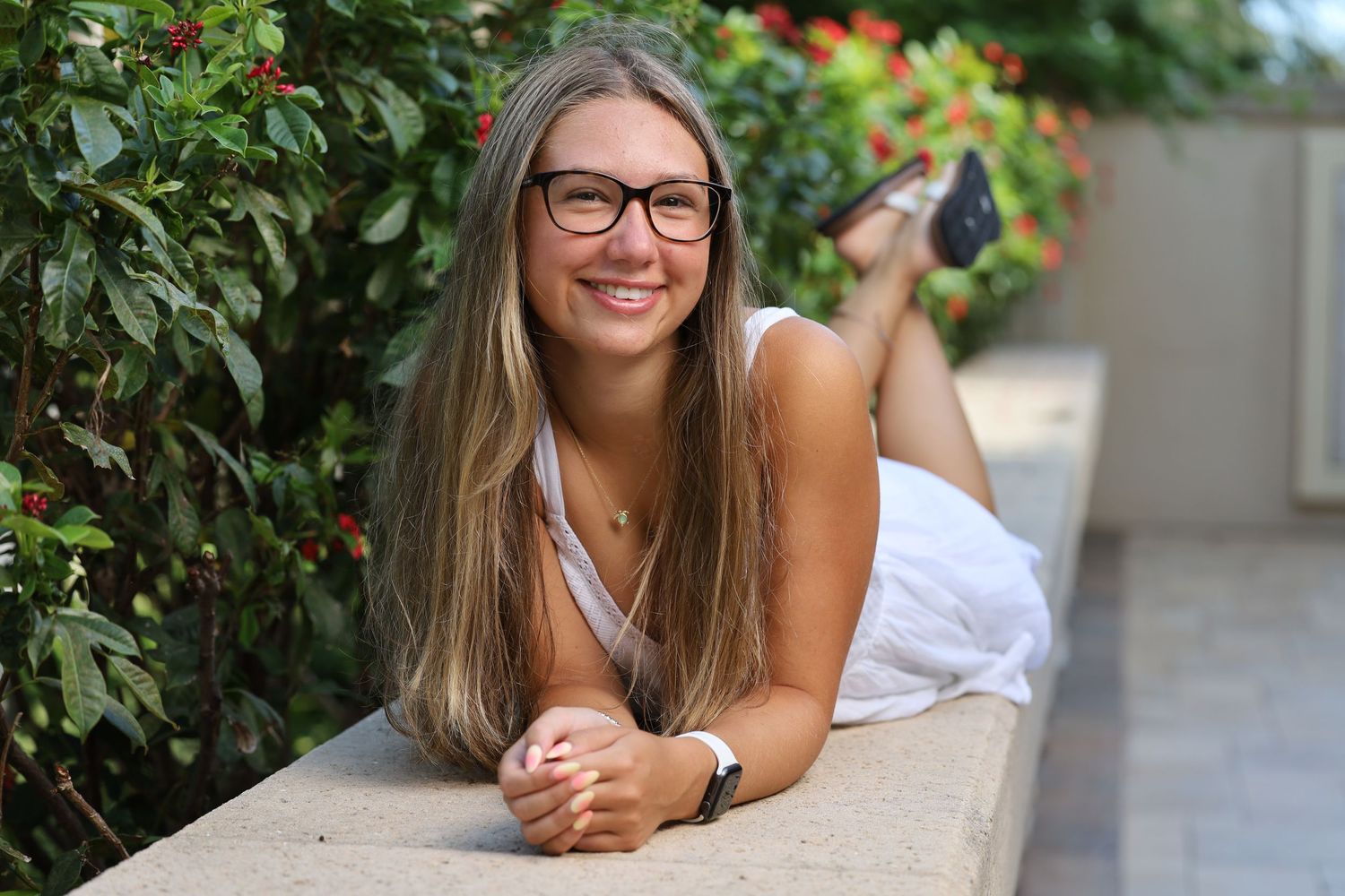 Smiling young woman with glasses and long blonde hair lying on an outdoor concrete ledge.