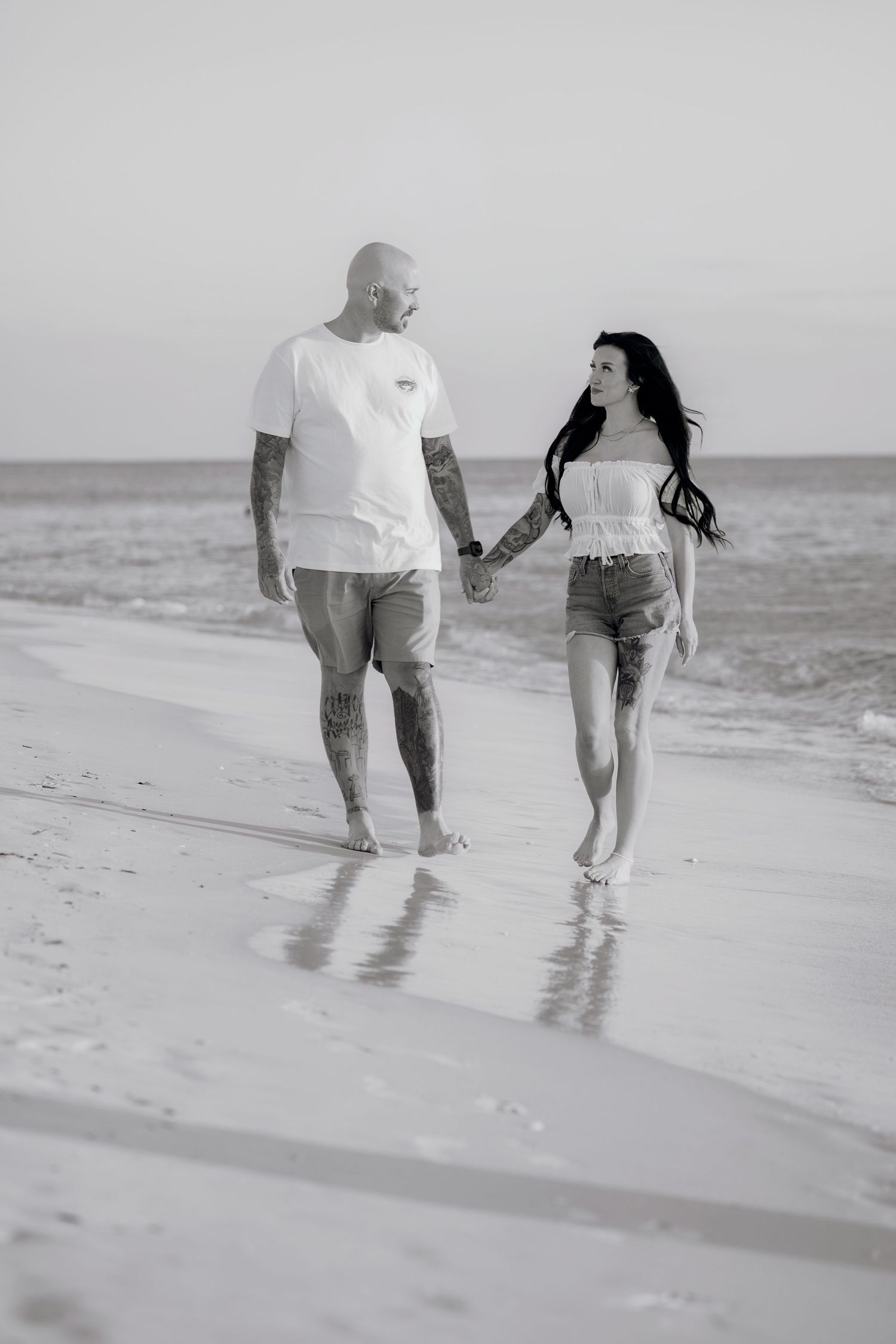 Black and white photo of a couple holding hands and walking away along a calm sandy shoreline