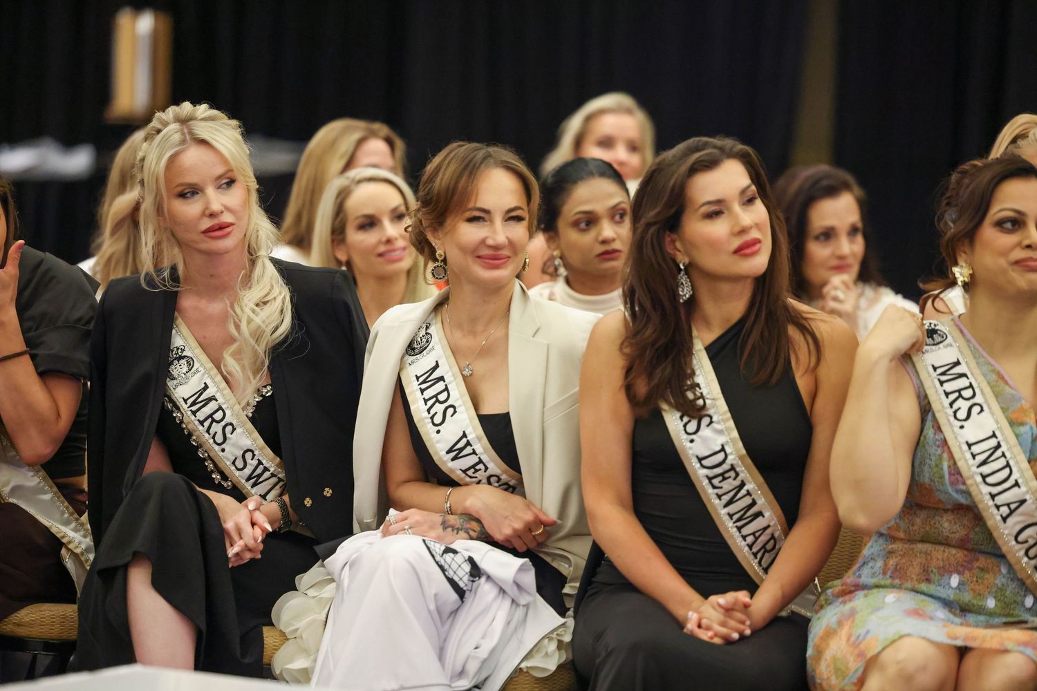 Pageant contestants sitting together, looking to the left while one contestant smiles confidently toward the camera lens