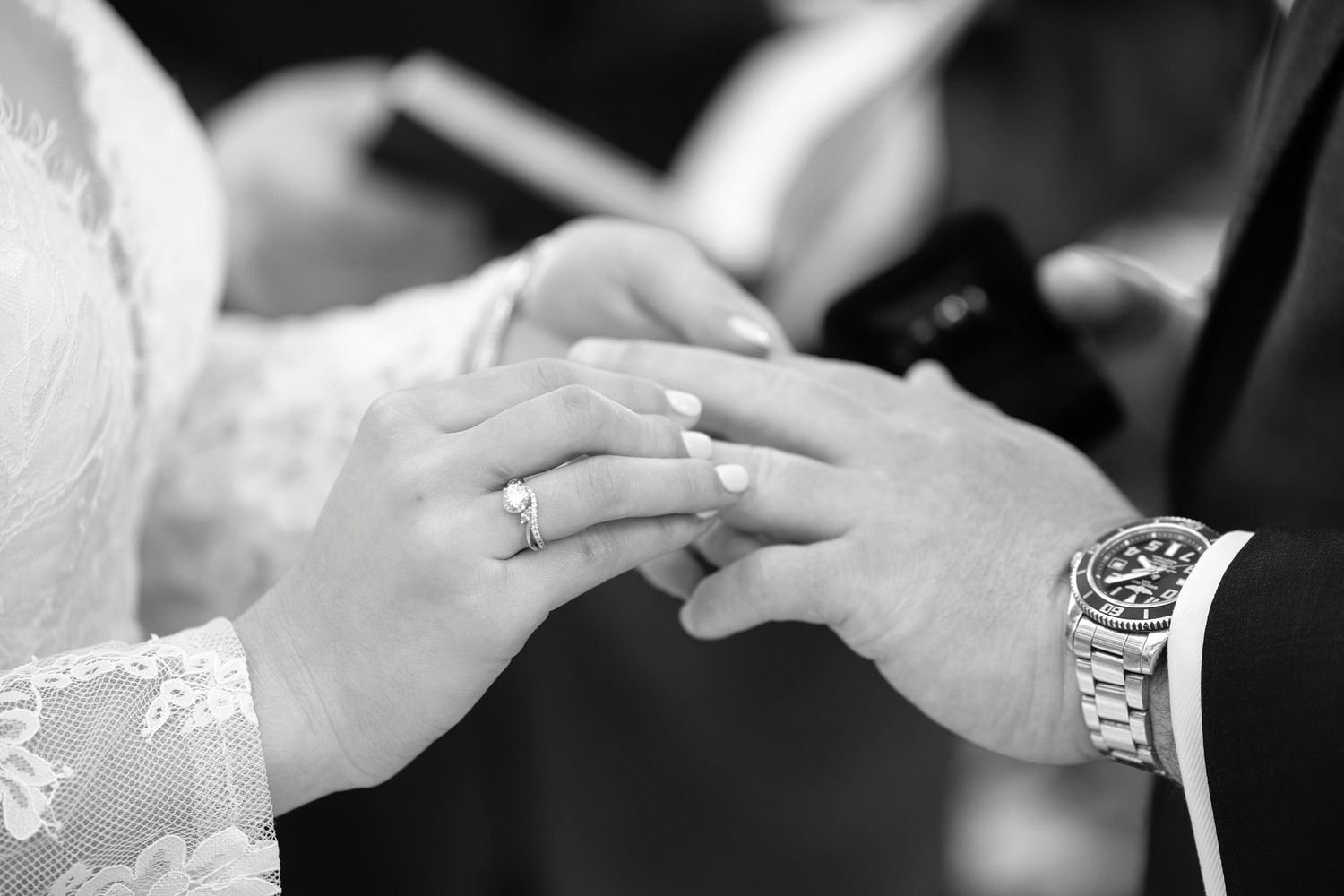 Black and white close-up of a bride placing a wedding band on the groom's finger during a marriage ceremony.