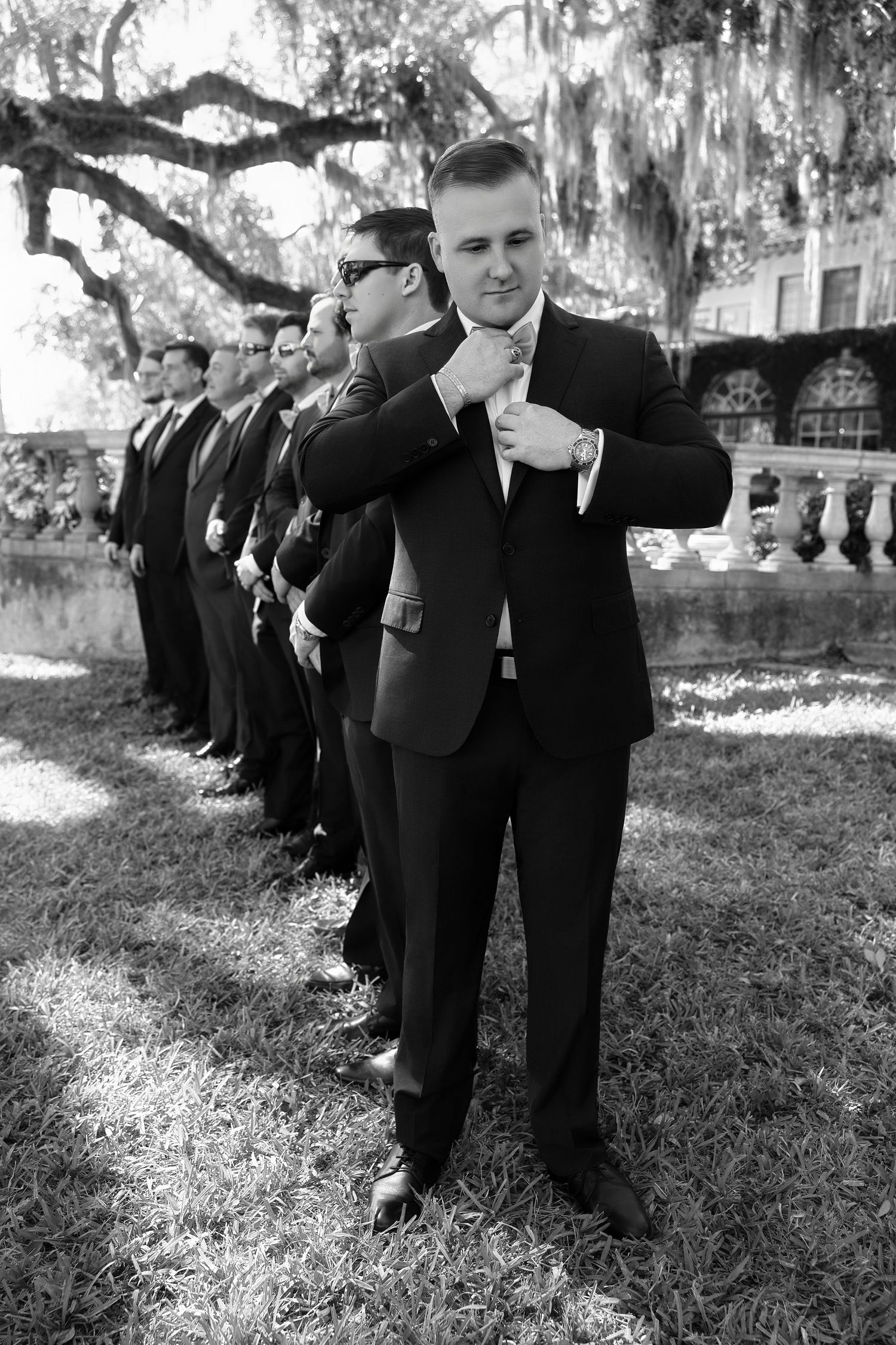 Black and white outdoor portrait of a groom standing in front of his groomsmen on a grassy lawn.