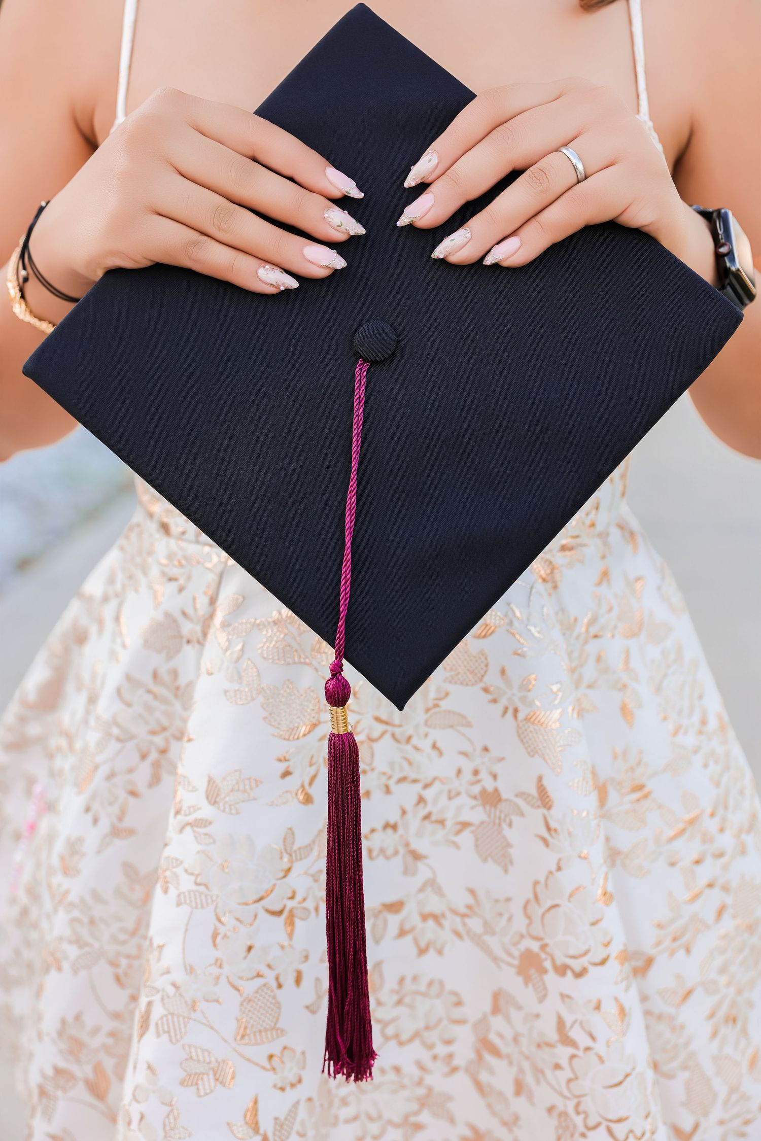Close-up of a graduate in a white lace dress holding a black mortarboard cap with a maroon tassel