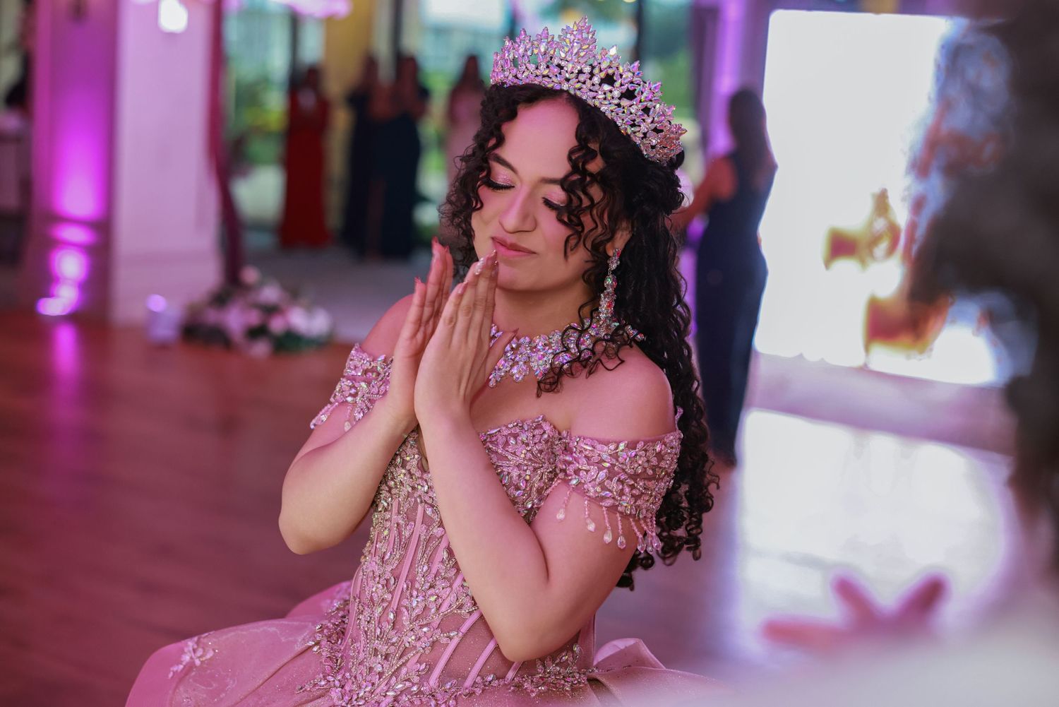 Young woman wearing a beaded pink gown and crown posing with hands together at a party venue