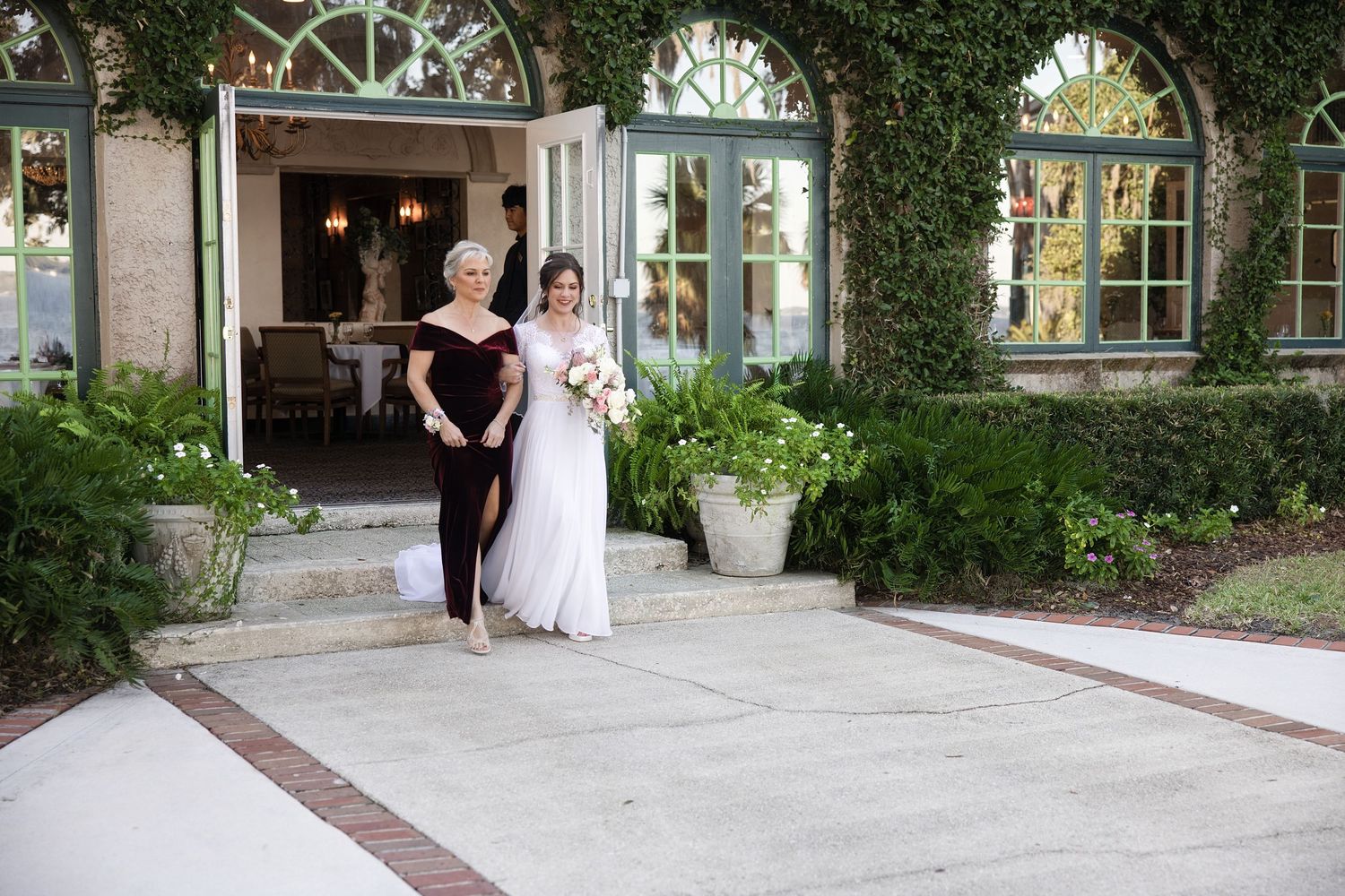 Bride walking down an outdoor paved pathway with an older family member outside a venue entrance