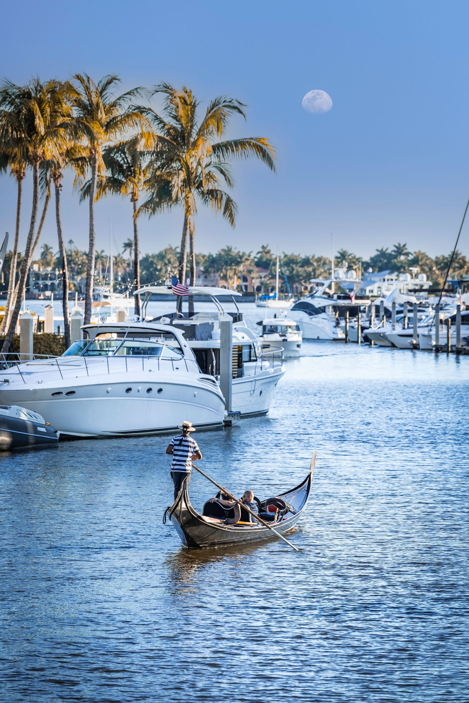 Traditional gondola boat navigating a coastal harbor lined with white luxury yachts and palm trees