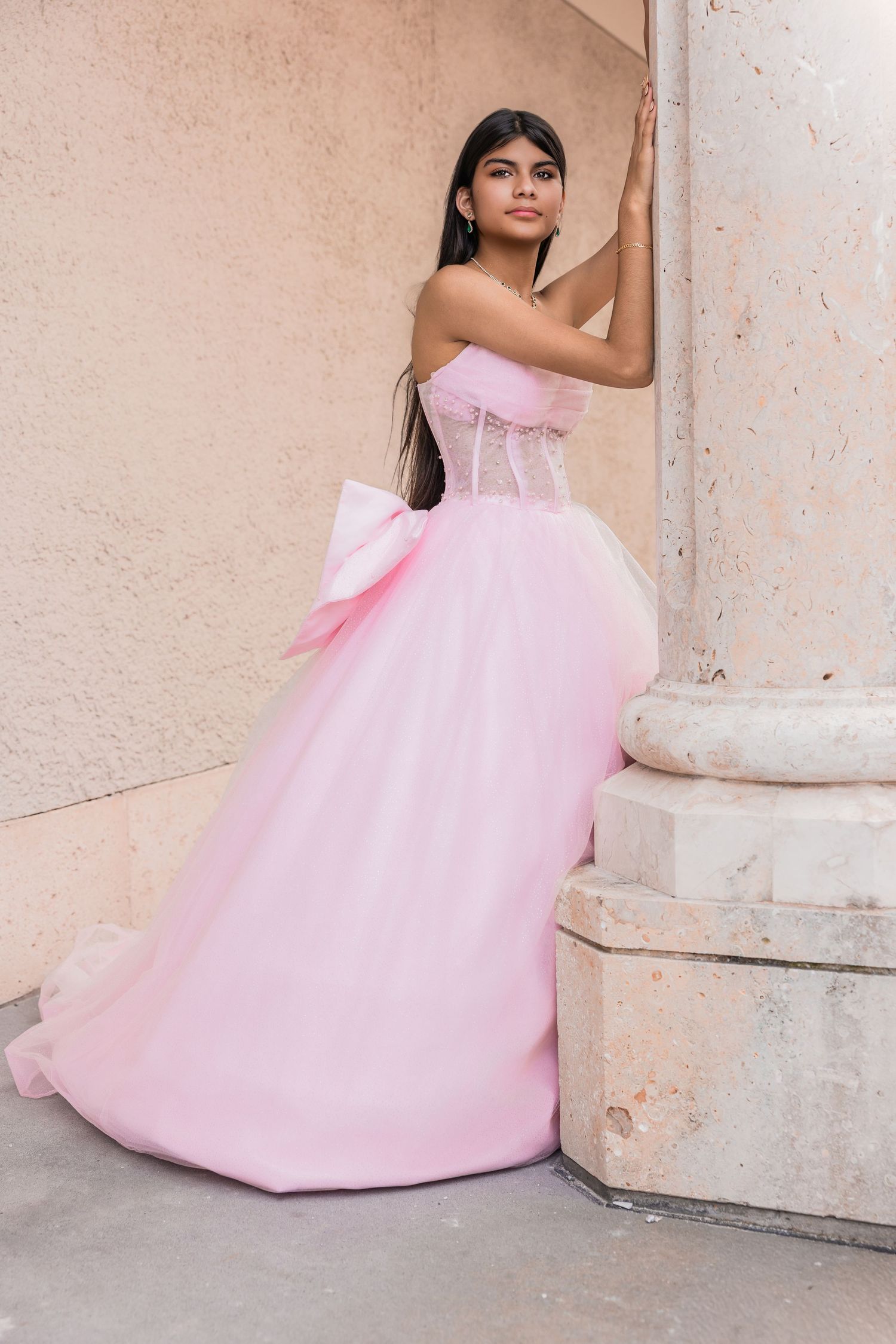 Young woman wearing a voluminous pink strapless ballgown leaning against a white column