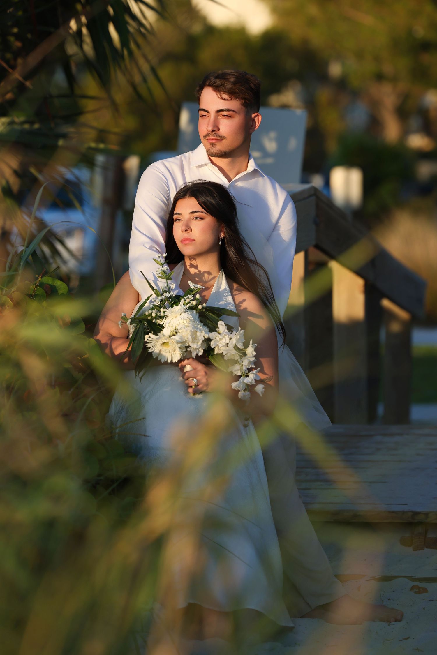 Groom embracing his bride from behind on a wooden garden bridge while she holds a wedding bouquet