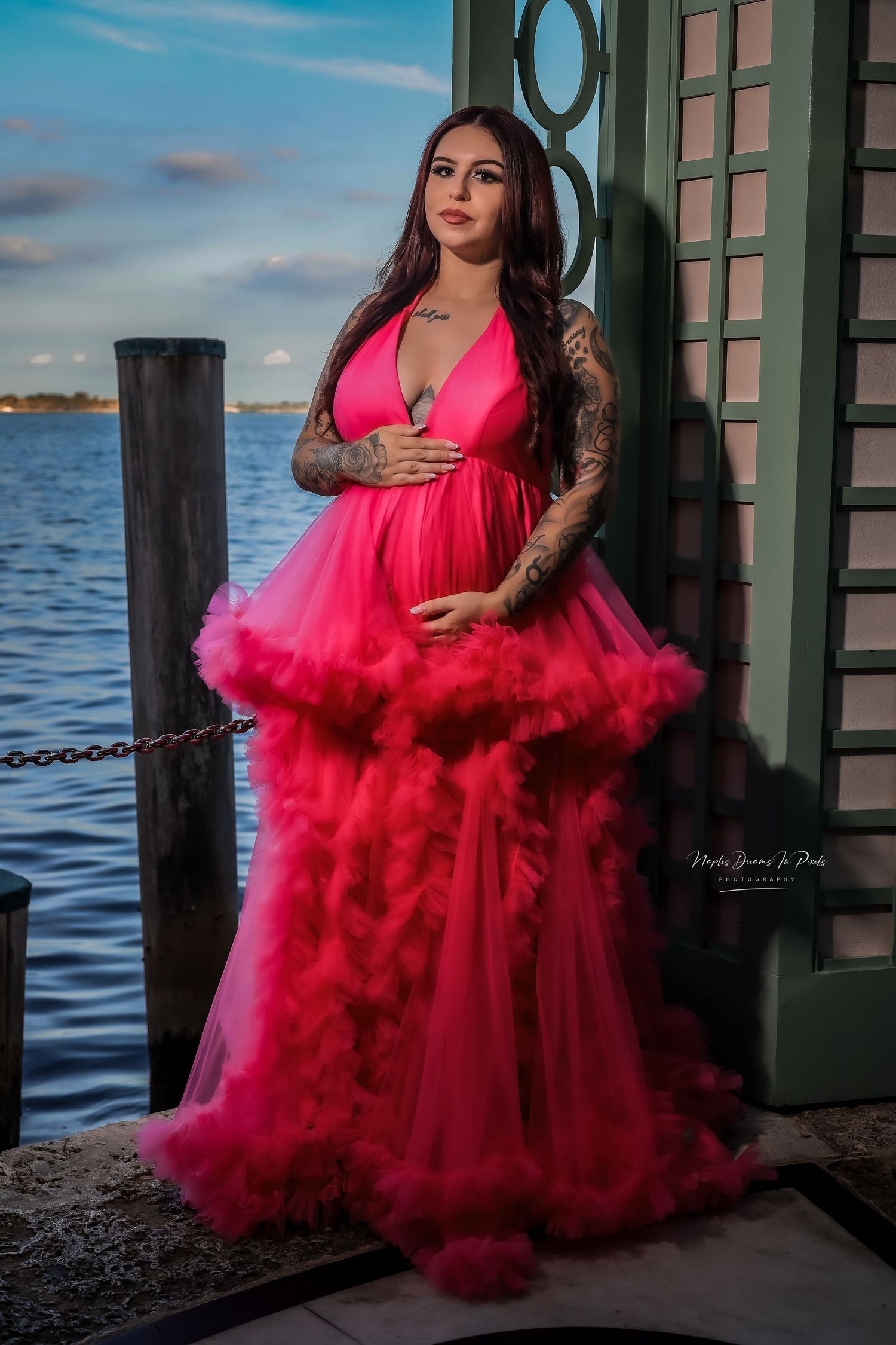 A pregnant woman poses by the water at sunset wearing a dramatic, multi-tiered hot pink tulle maternity dress.