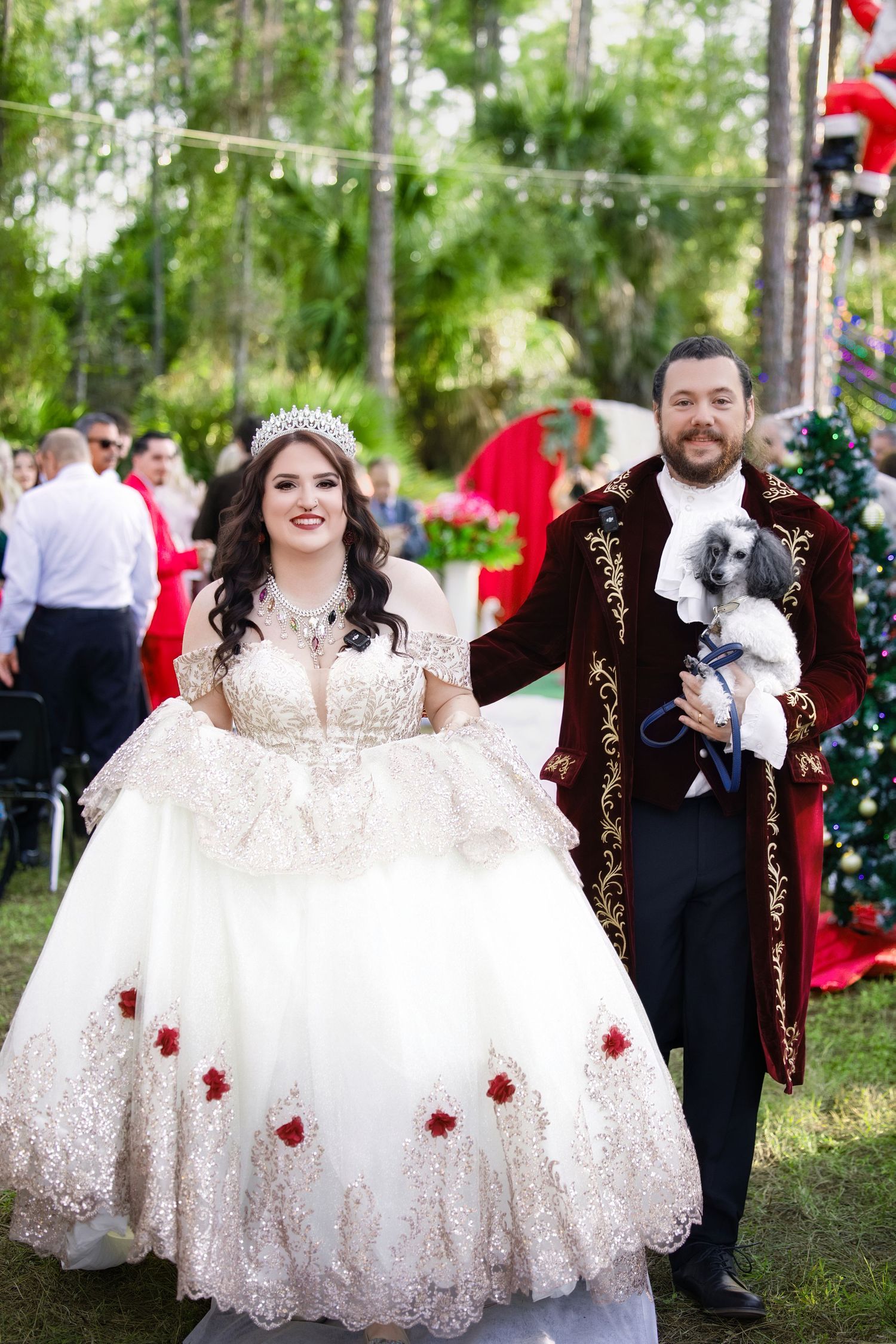 Bride in a floral embellished ballgown and groom in a red velvet coat walking down the outdoor ceremony aisle