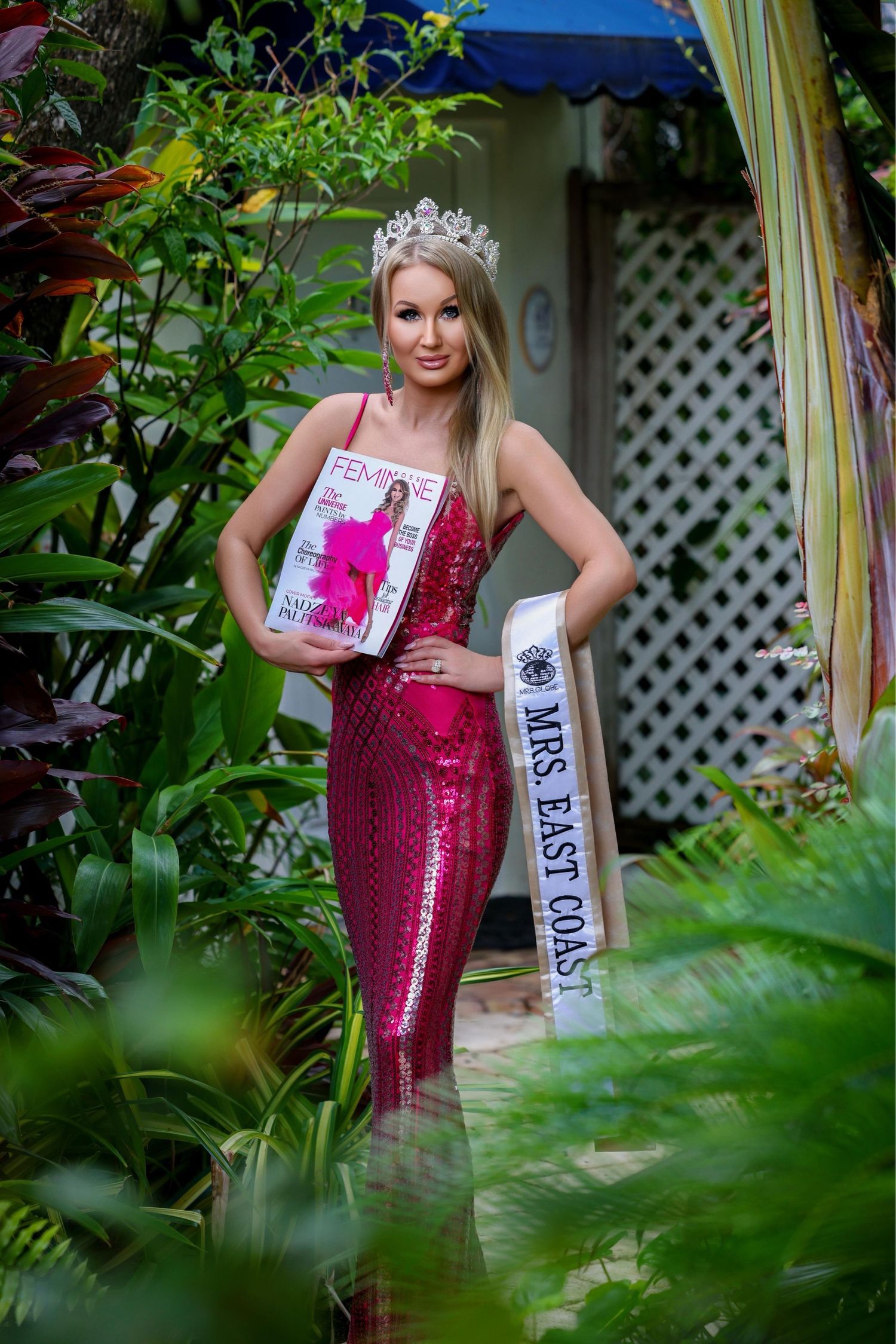 Pageant winner wearing a magenta dress, crown, and sash posing in a tropical garden setting