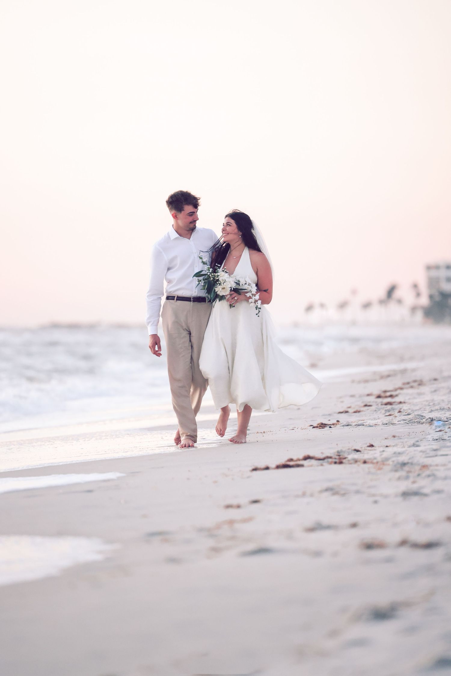 Newlywed bride and groom walking barefoot along the ocean tide line on a beach at sunset