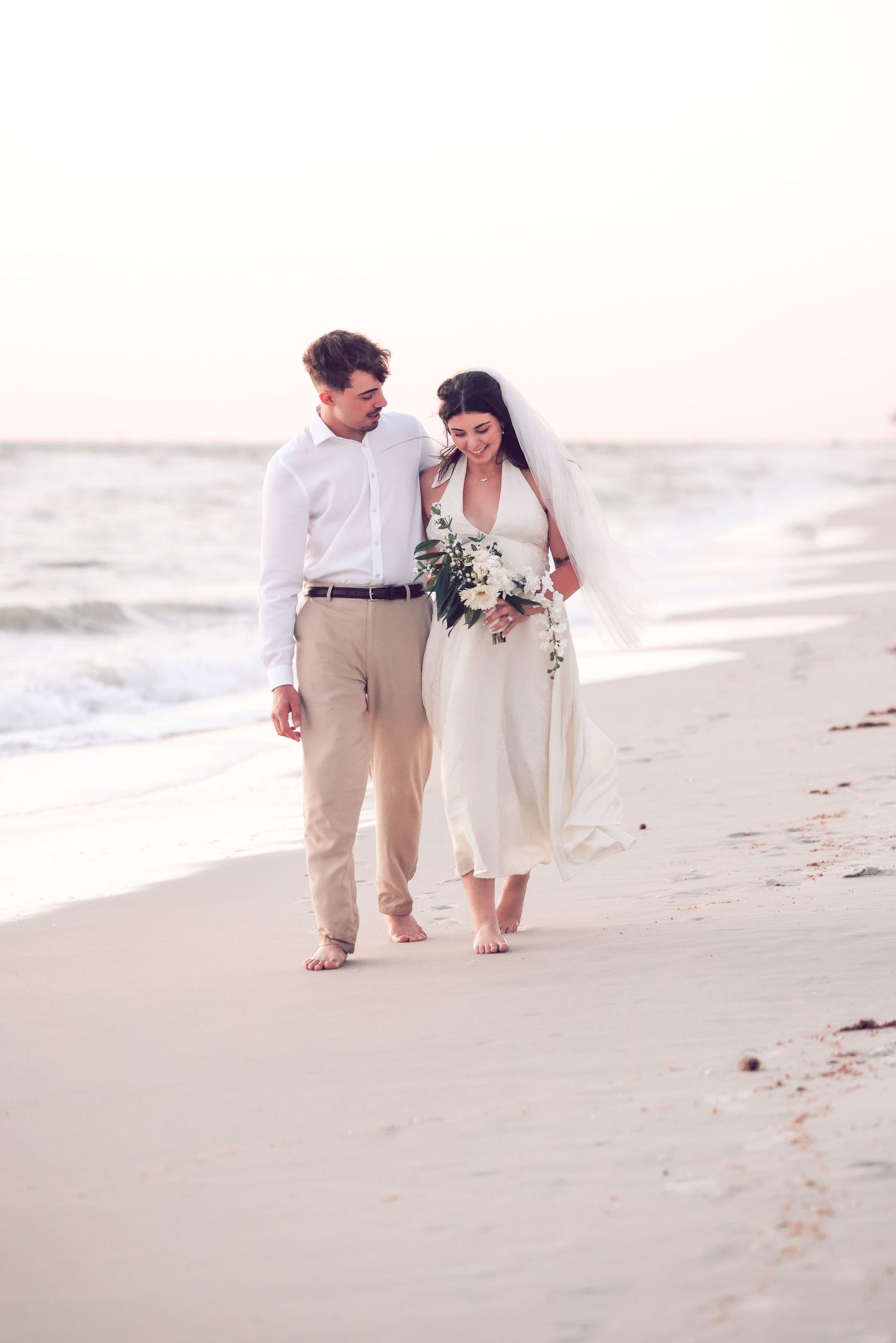 Bride and groom walking barefoot hand-in-hand along the sandy shore during a beach wedding at sunset.