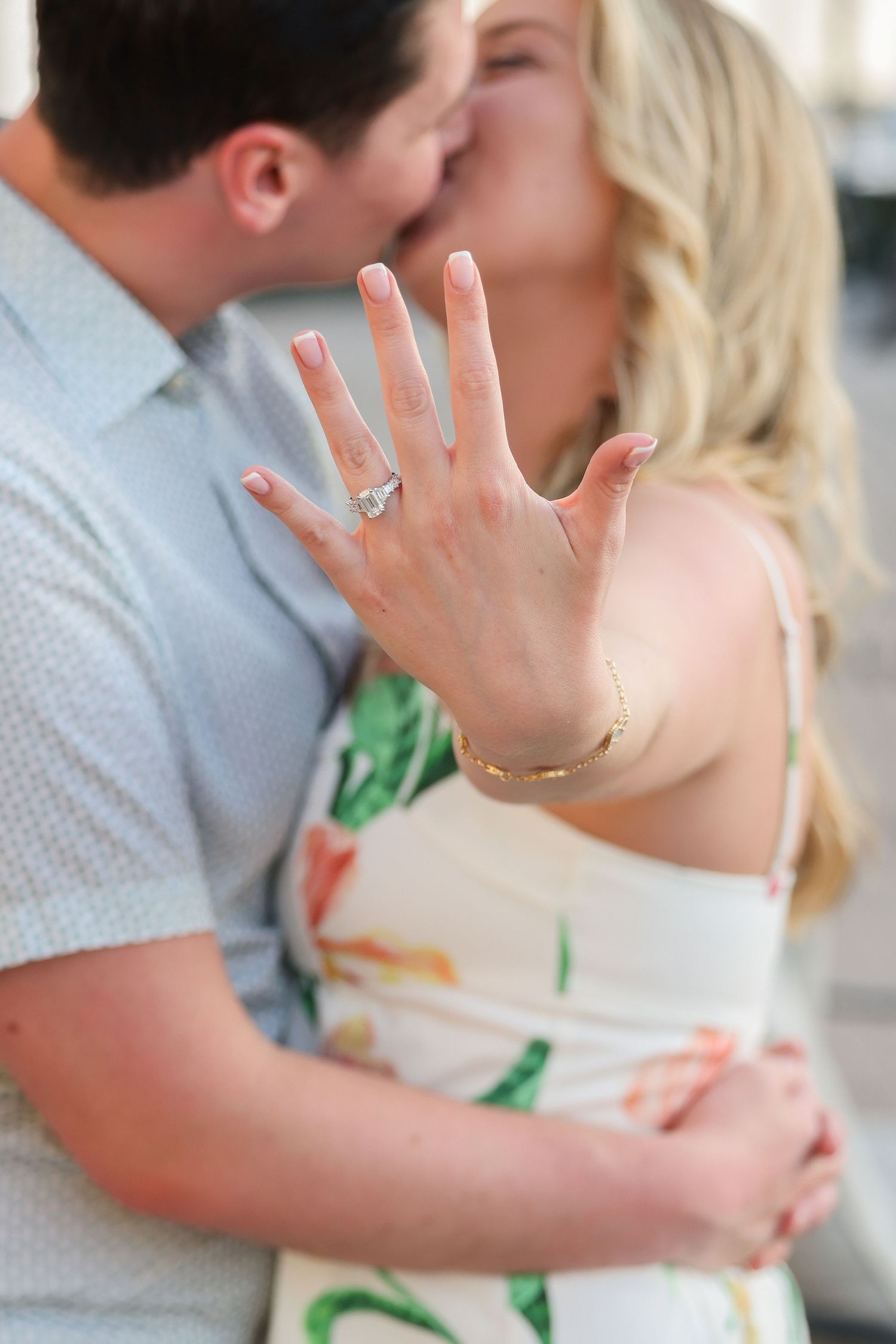 Close-up of a blonde woman showing her diamond engagement ring while kissing her fiancé in the background