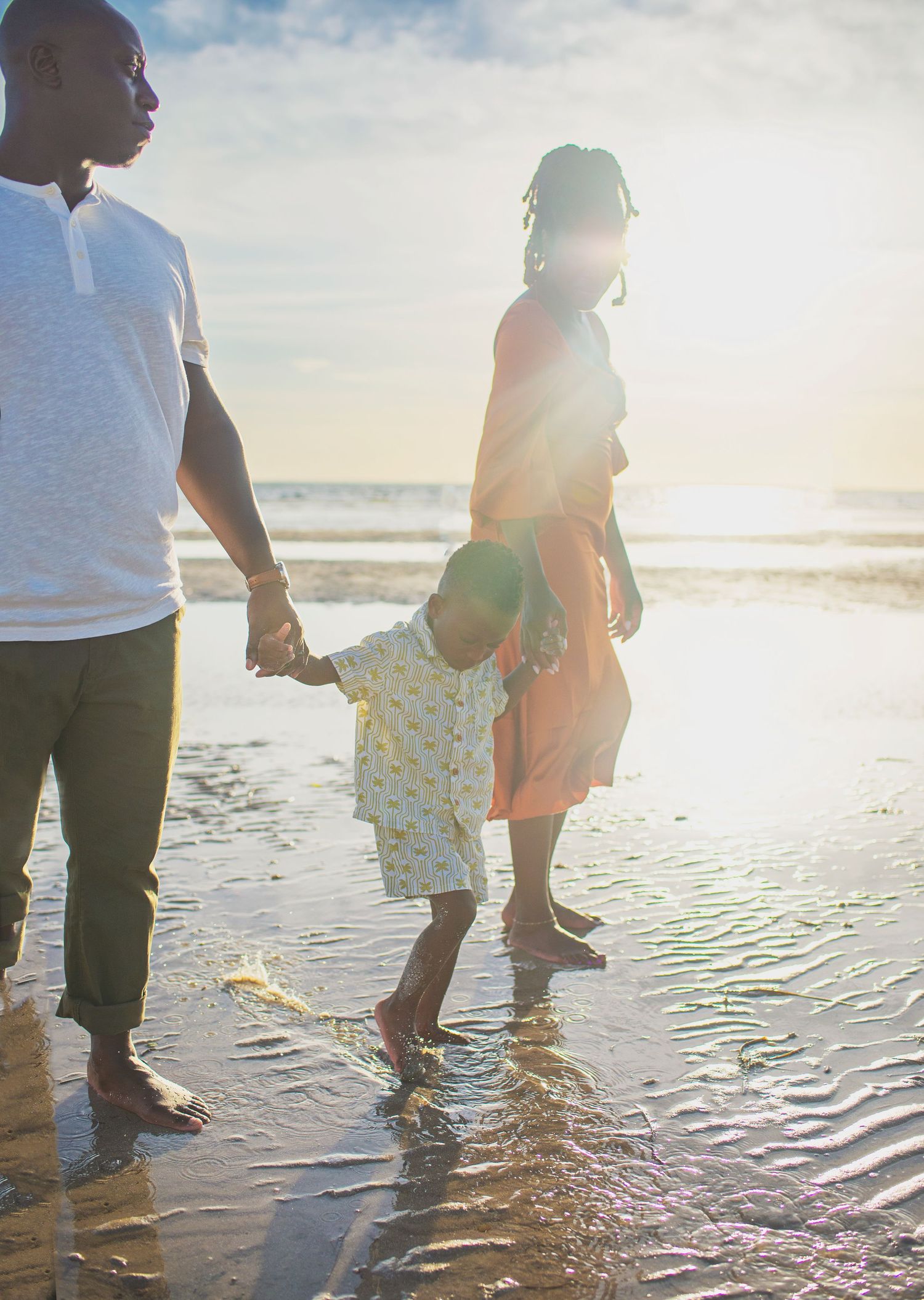 A family of three holds hands while wading in shallow beach water at sunset.