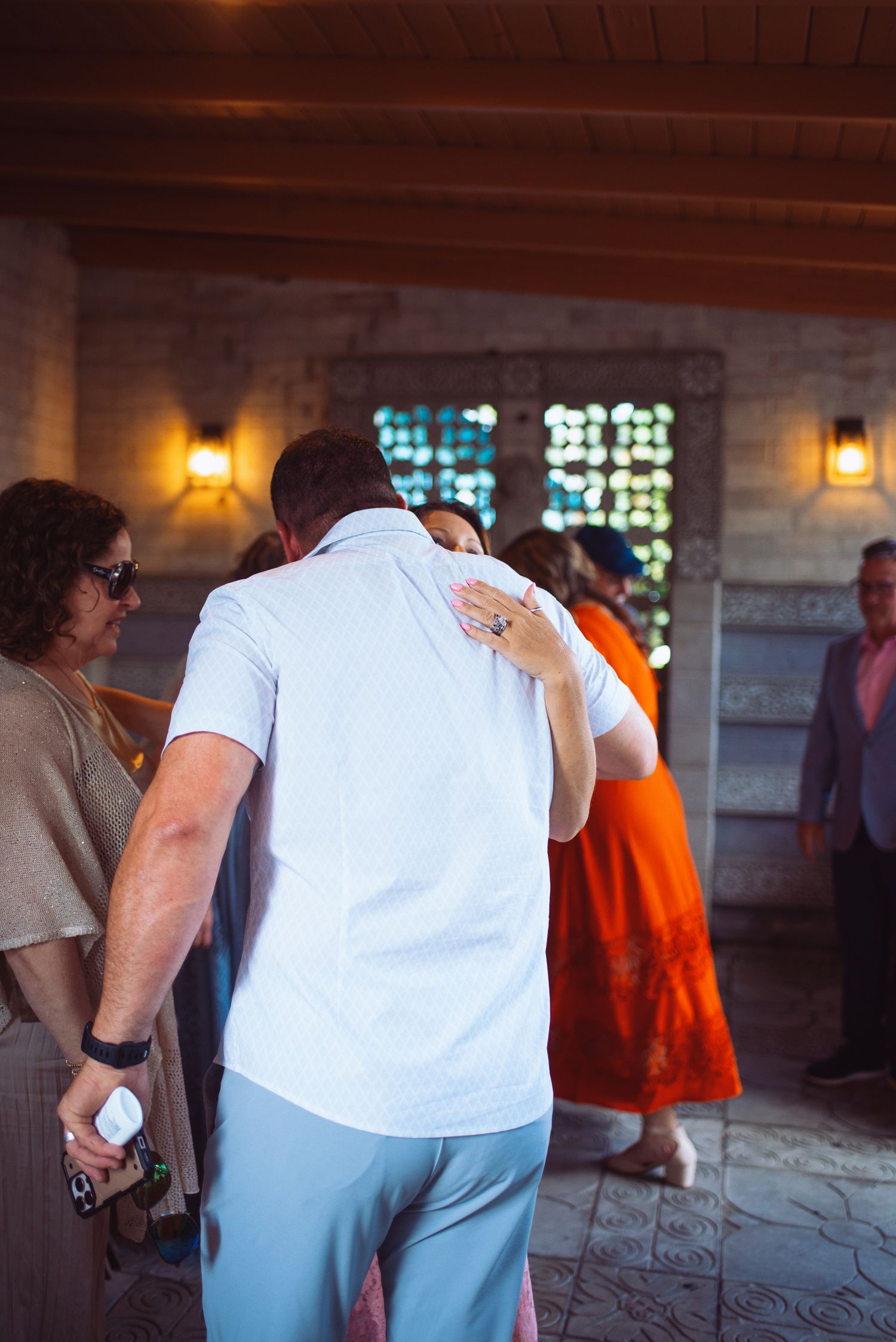 Wedding guests mingle and embrace inside a warmly lit stone venue with wooden beam ceiling.