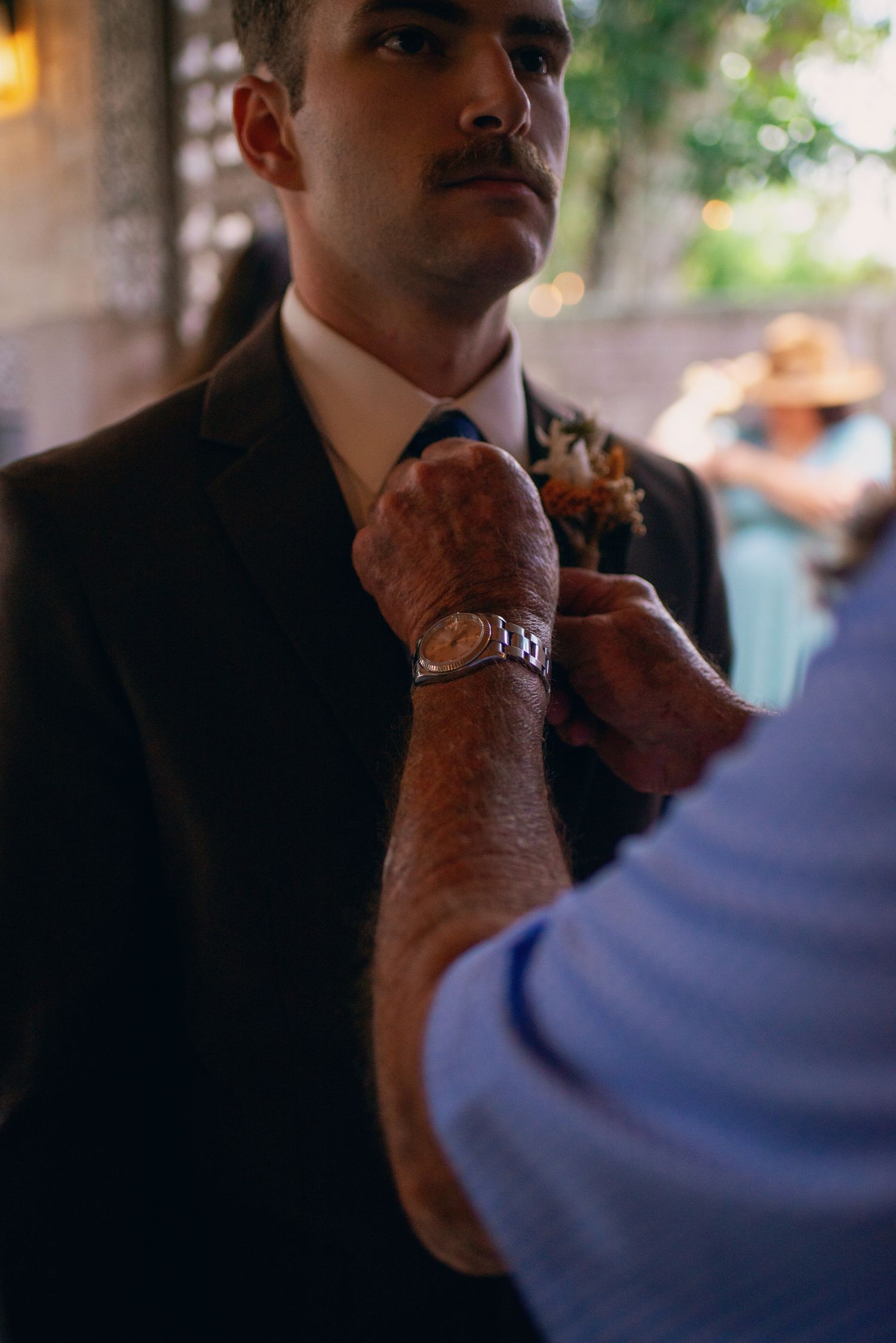 Elder hands pin floral boutonniere on groom in dark suit with mustache at wedding.