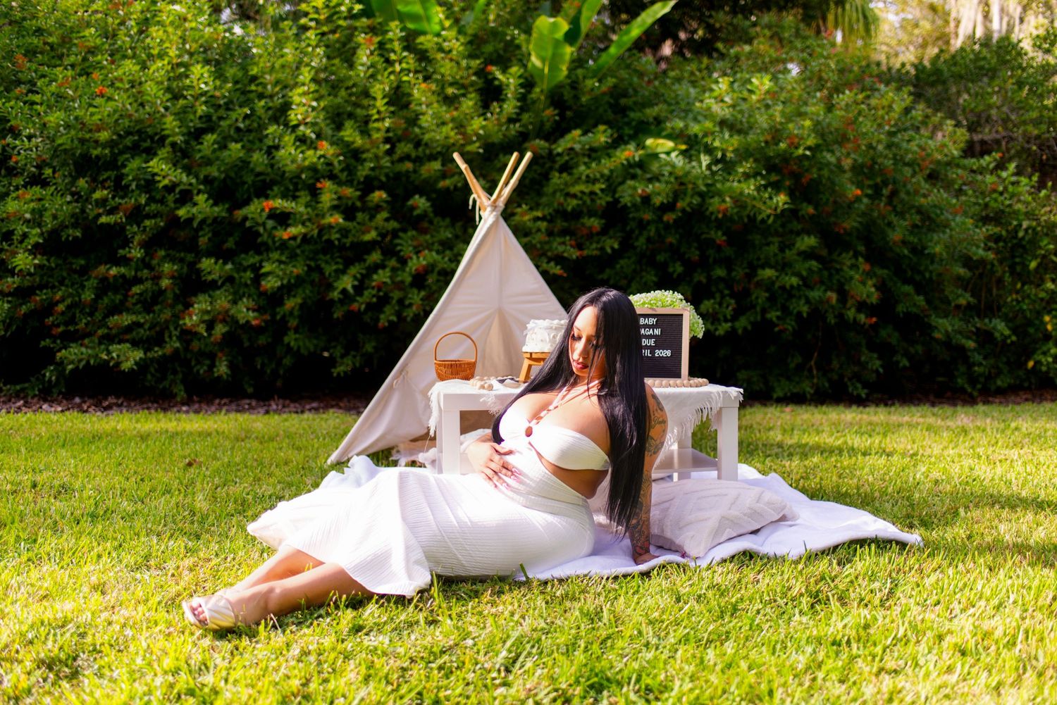 Woman in a white dress relaxing on a blanket beside a teepee setup on a lush green lawn.