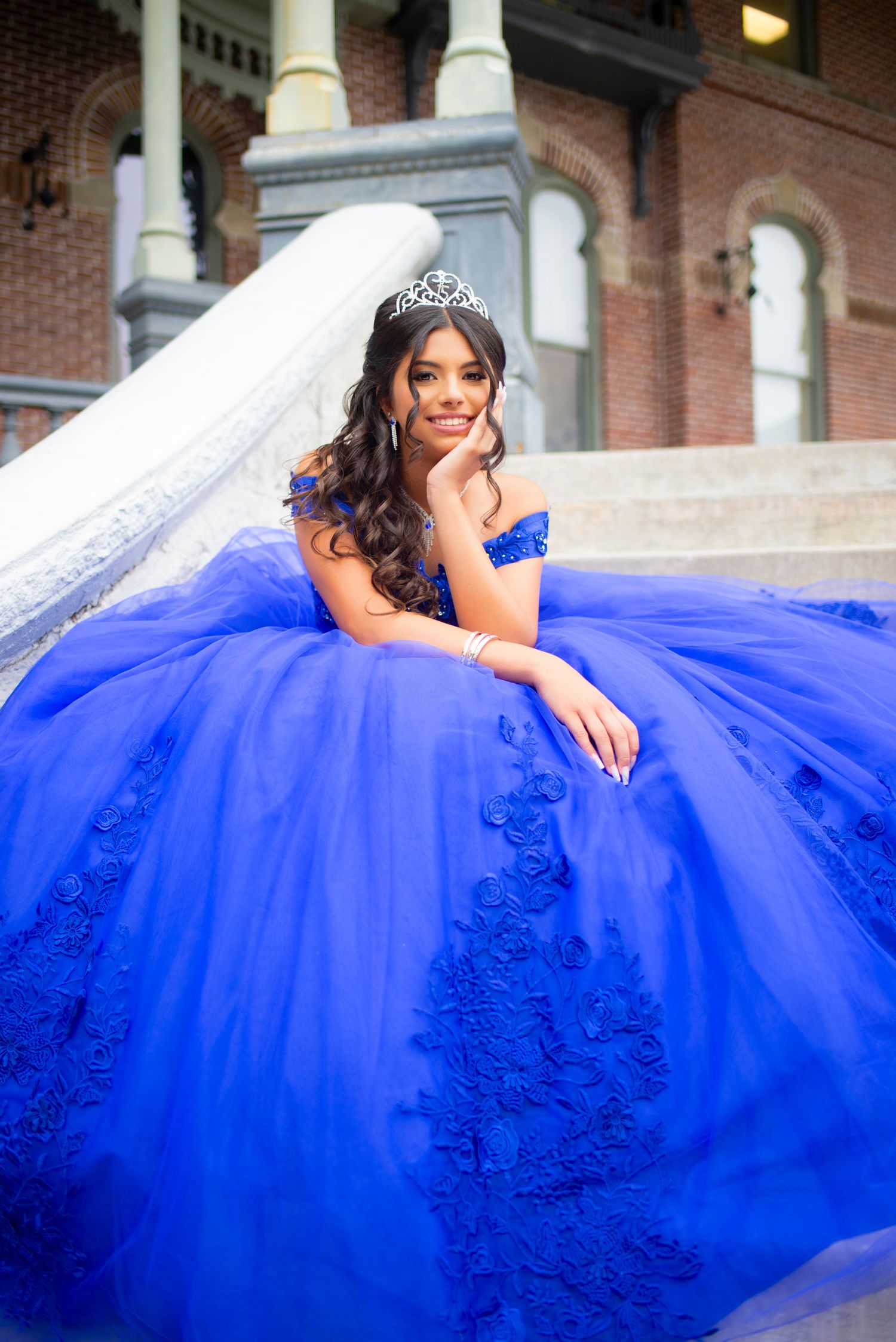 Quinceañera girl in royal blue lace ball gown and tiara posing elegantly on steps of a historic building.
