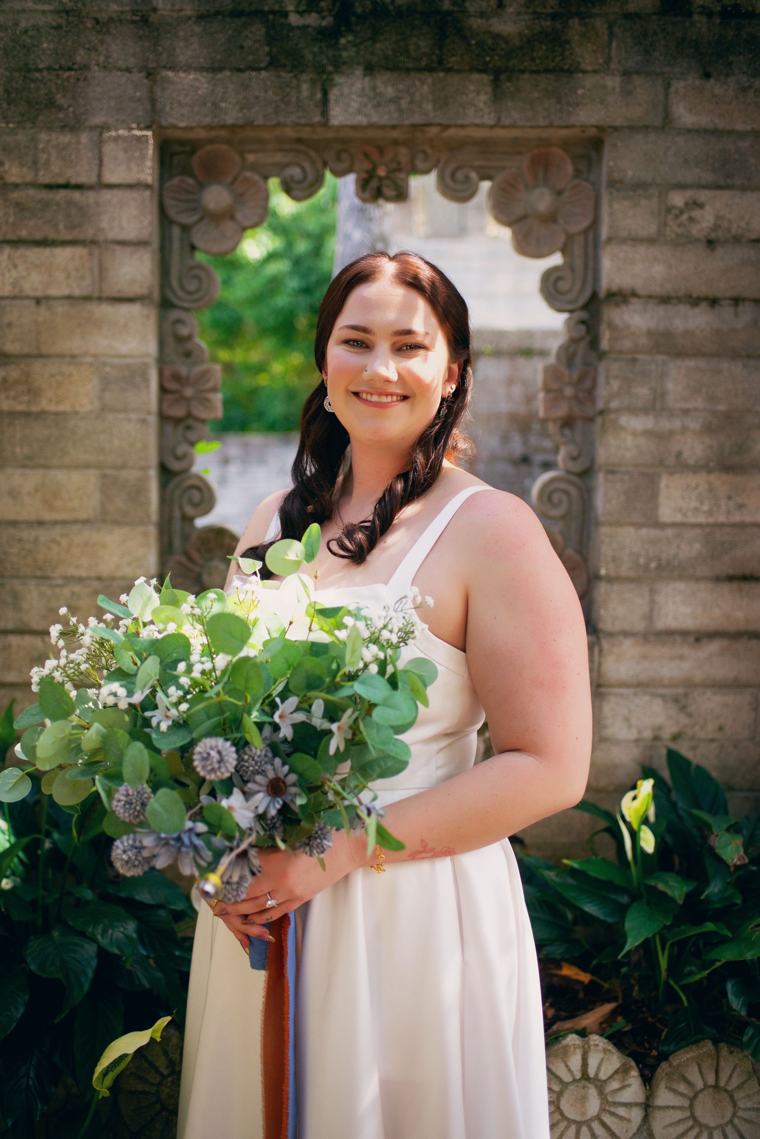 Bride smiling in white dress holding eucalyptus bouquet beside ornate stone wall garden feature.