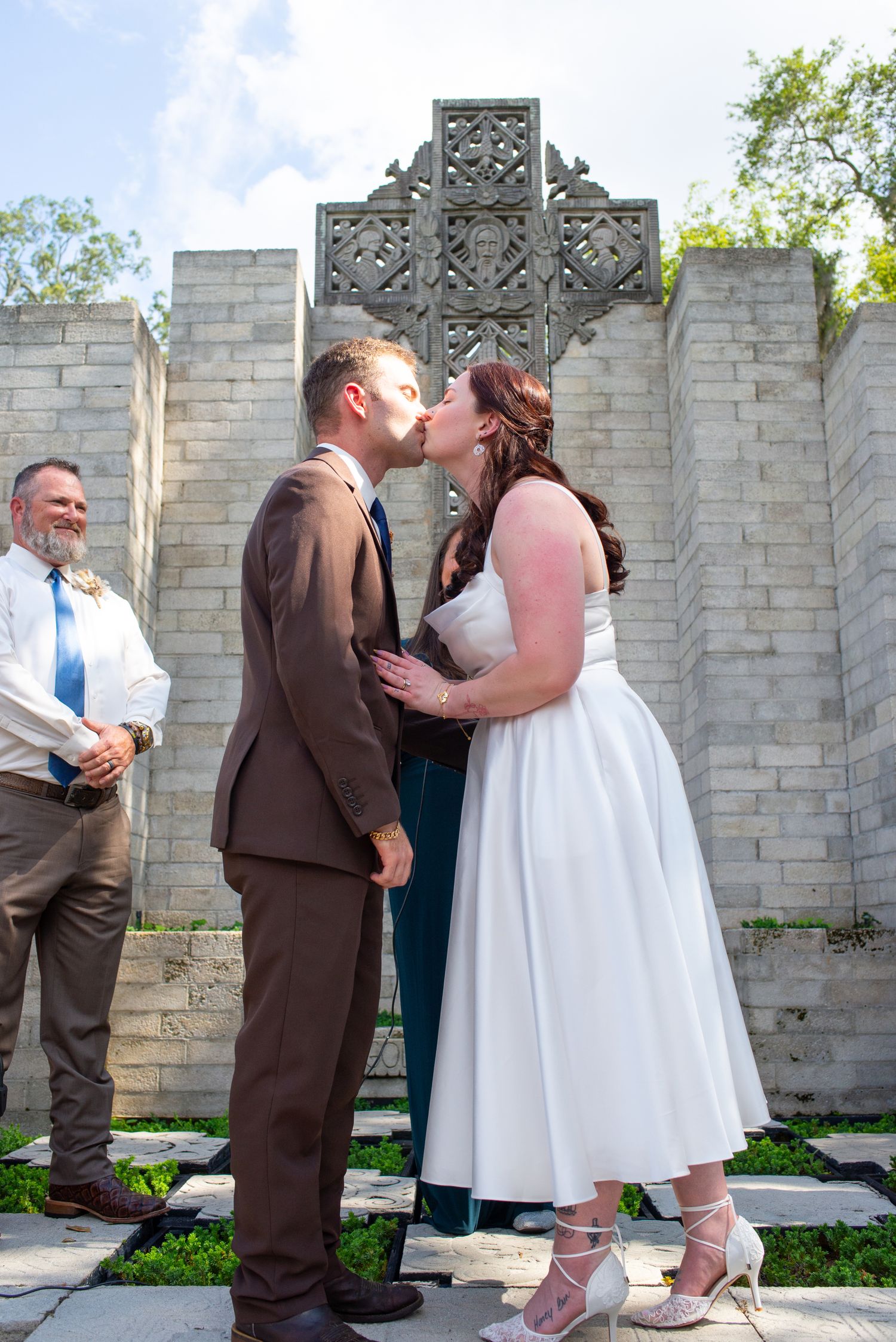 A bride and groom share their first kiss in front of a stone cross at an outdoor wedding.