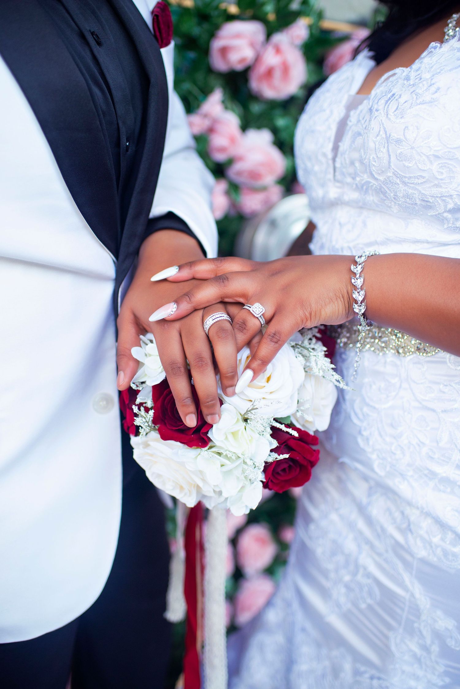 Bride and groom display wedding rings while holding red and white rose bouquet together.