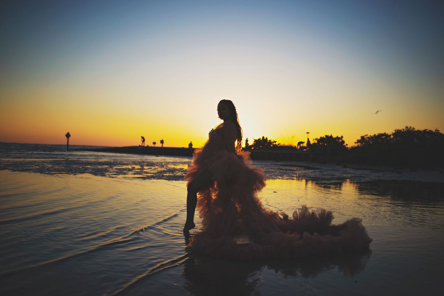 Silhouette of a woman in a ruffled gown standing in shallow water during a vibrant sunset.