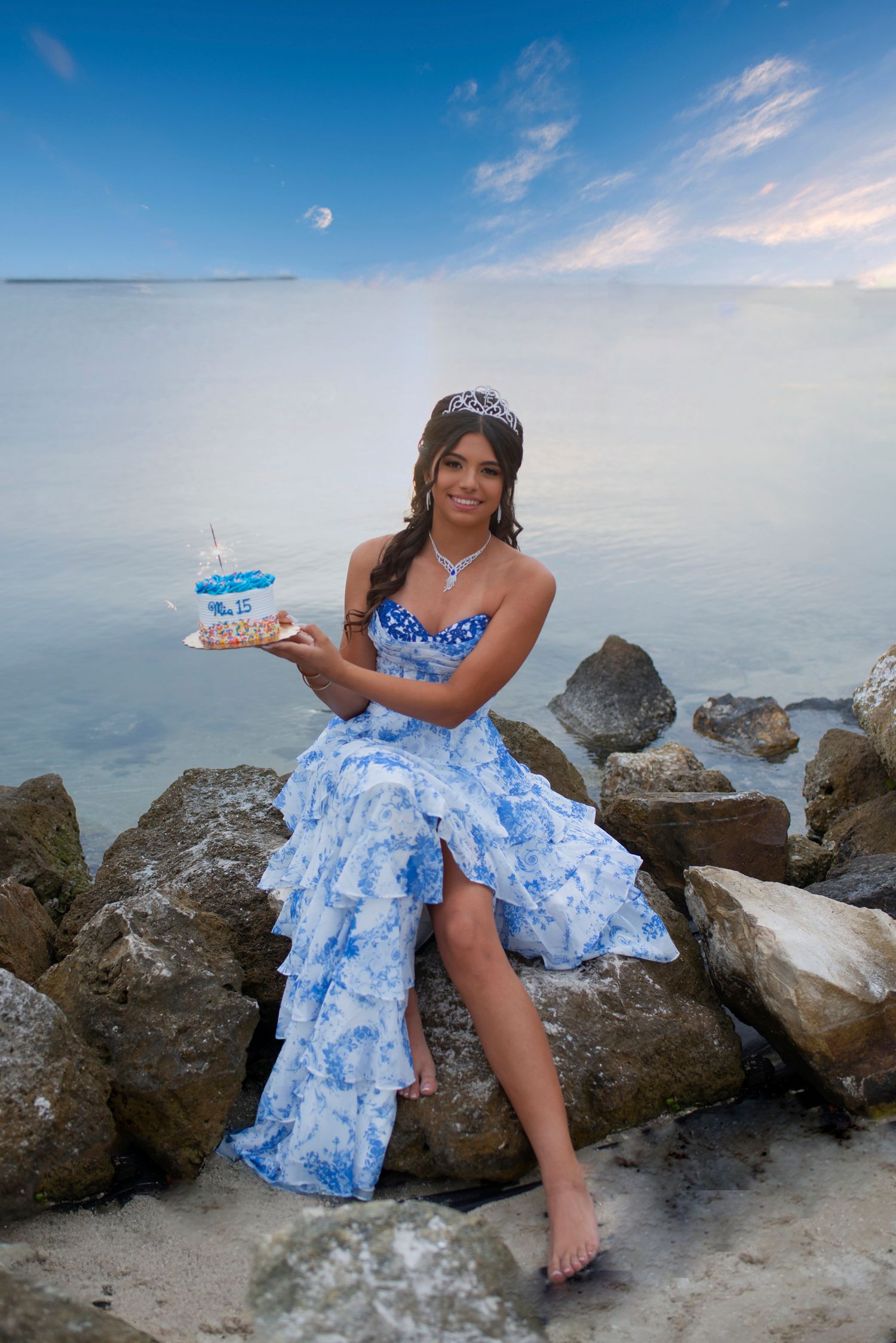 A smiling young woman in a blue floral dress holds a birthday cake while sitting on rocks by the sea.