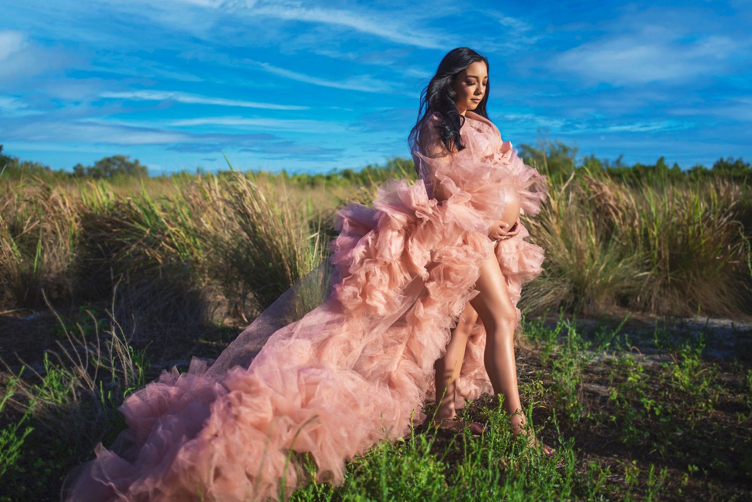 Pregnant woman in flowing pink ruffled maternity gown posing outdoors in grassy field with blue sky.