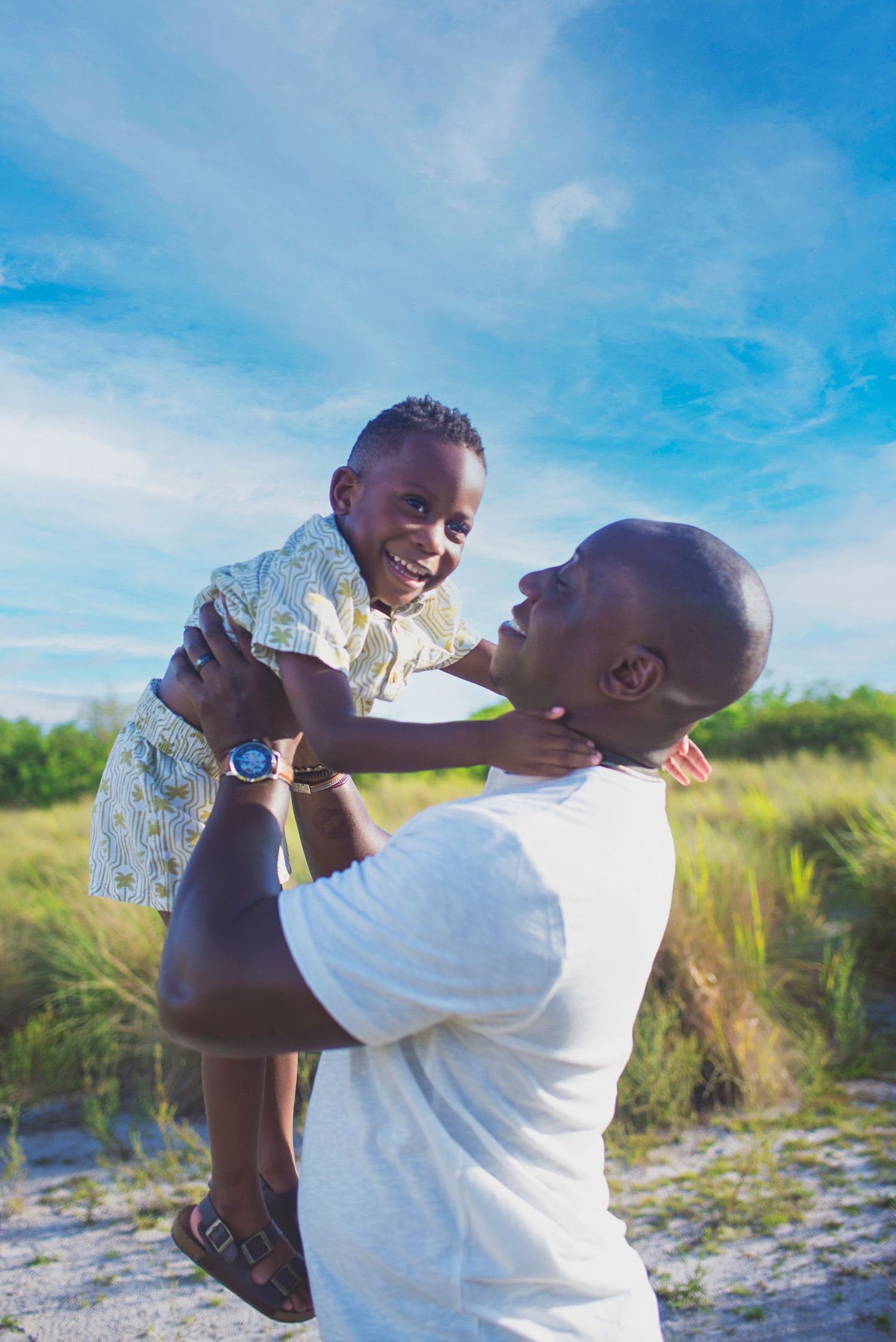 A joyful father lifts his laughing young son against a bright blue sky outdoors.