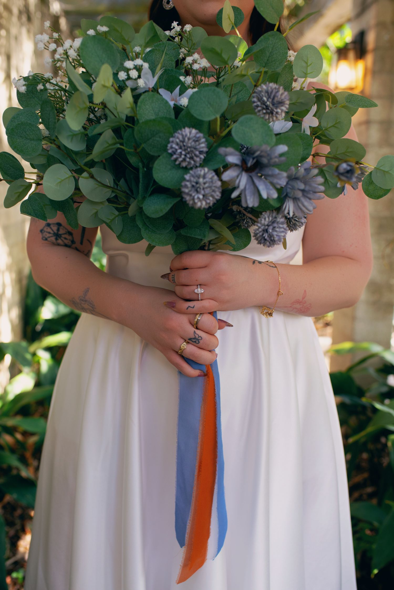 Close-up of bride holding wedding bouquet with blue ribbons, rings and bracelet visible on hands.