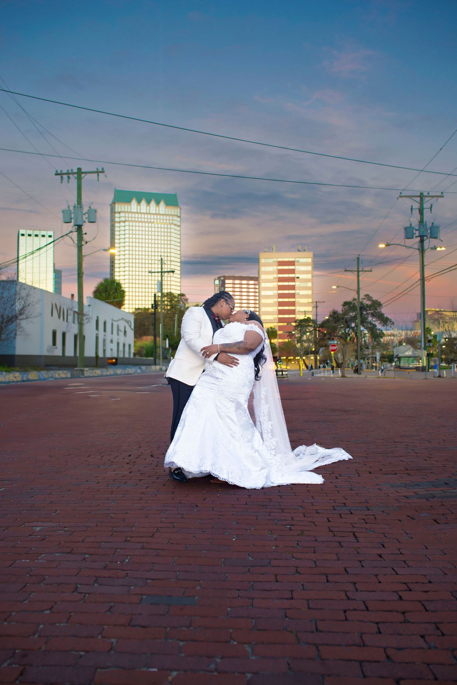 Wedding couple shares romantic dip kiss on brick plaza with city skyline at sunset.