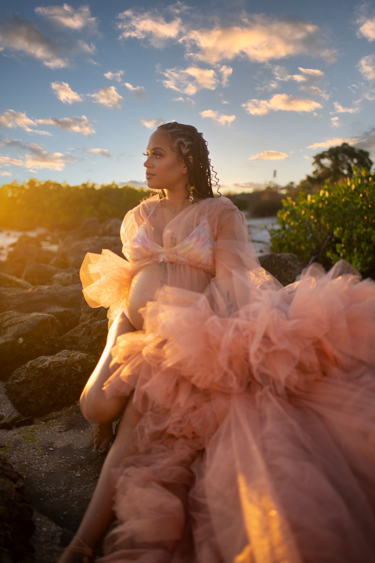 Pregnant woman in pink ruffled tulle gown sitting on rocks at glowing golden sunset.