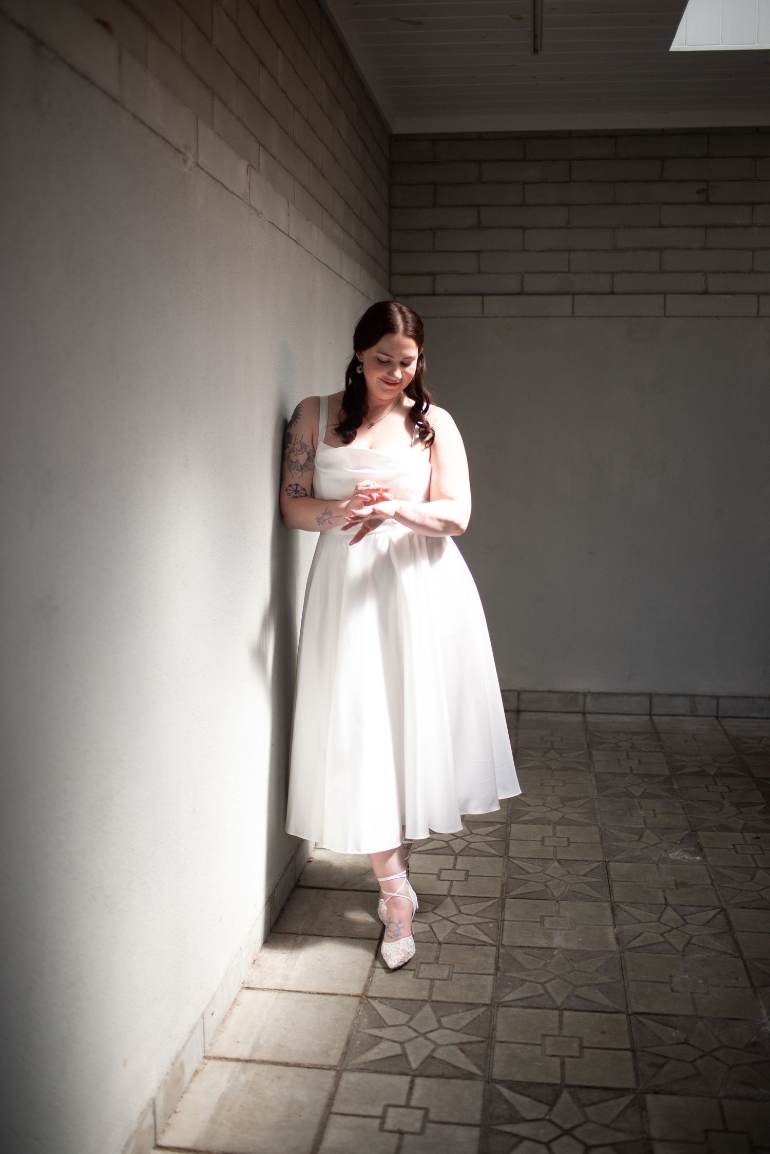 Bride in white midi dress leaning against sunlit wall in tiled corridor, looking down gracefully.