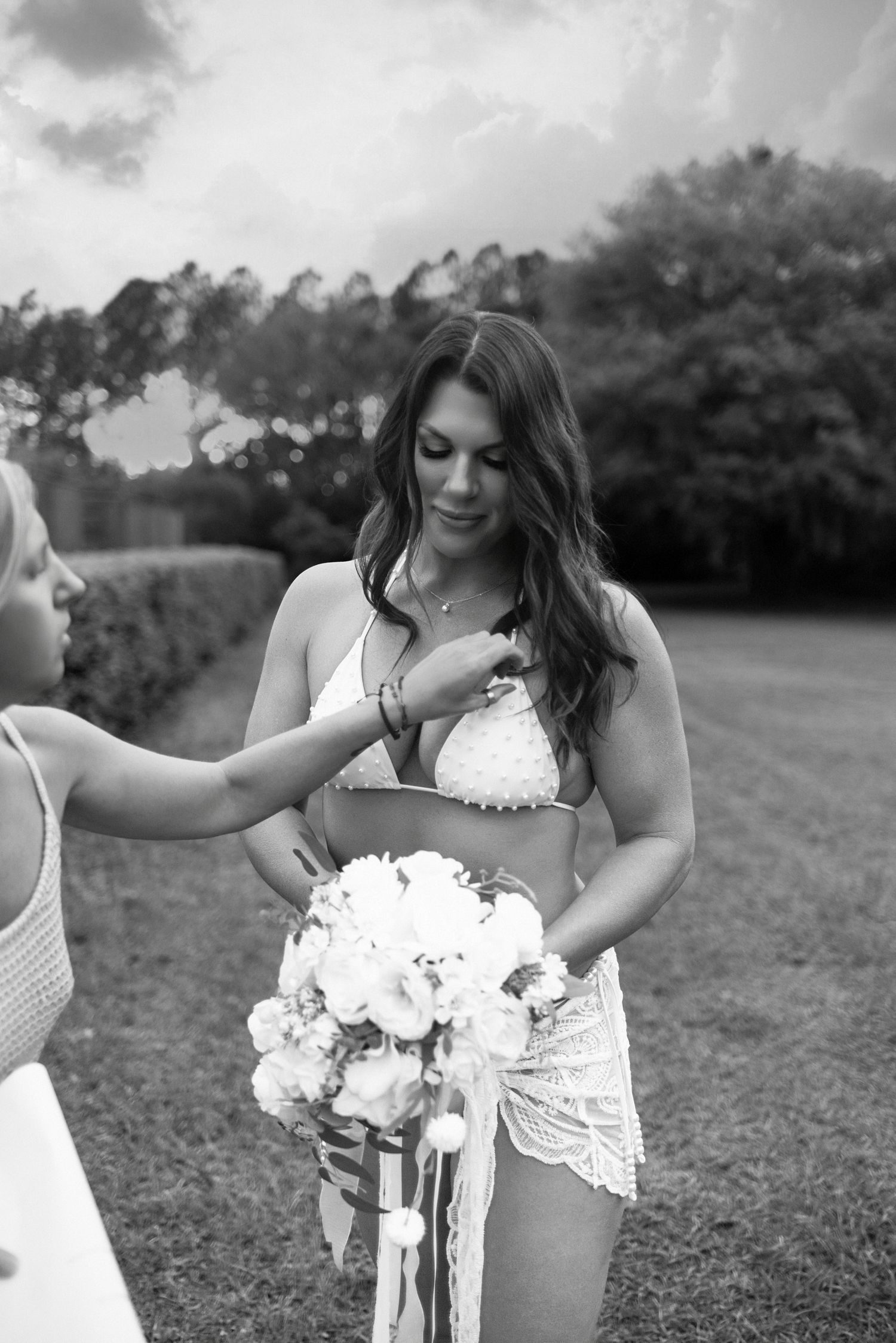 Black and white photo of bride in white bikini holding bouquet while friend adjusts her necklace outdoors.
