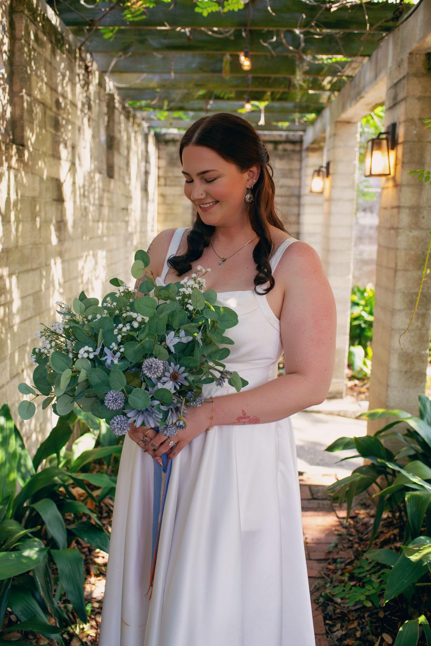 Bride in white dress gazes down at eucalyptus and blue thistle bouquet in sunlit garden corridor.