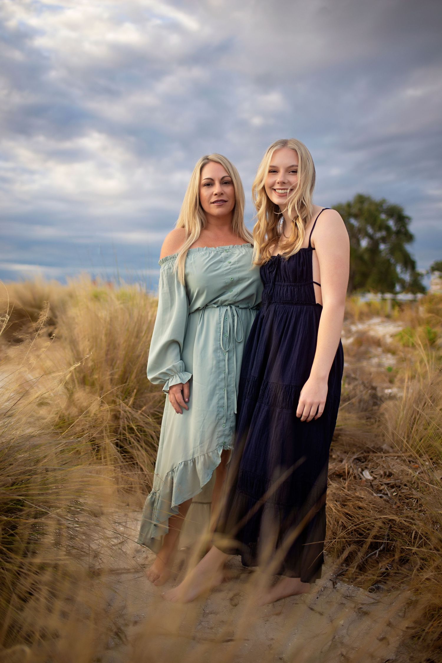 Two blonde women in elegant dresses pose together on a sandy beach at dusk.