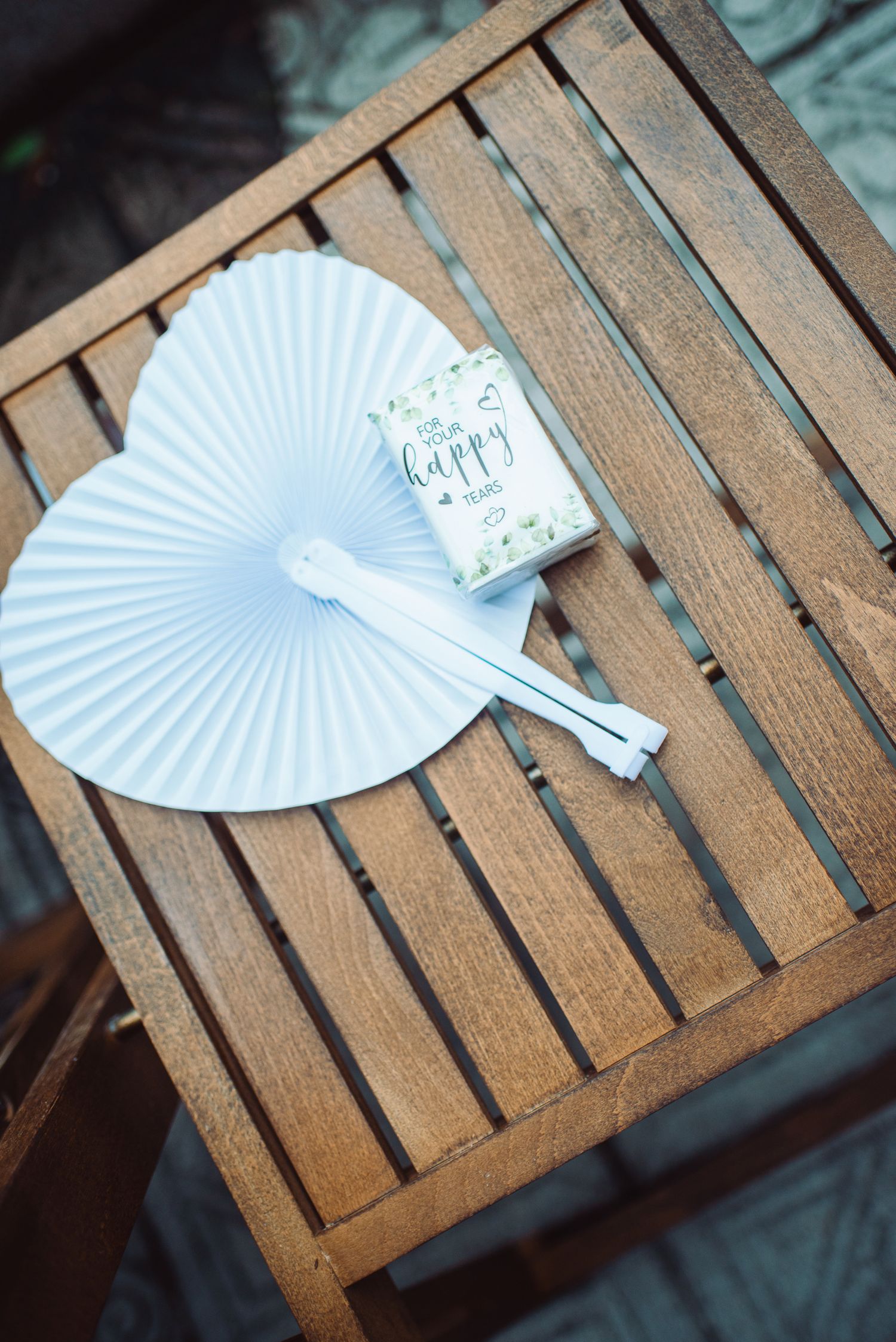 White paper hand fan and happy couple wedding favor box on a wooden slatted table at an outdoor event.